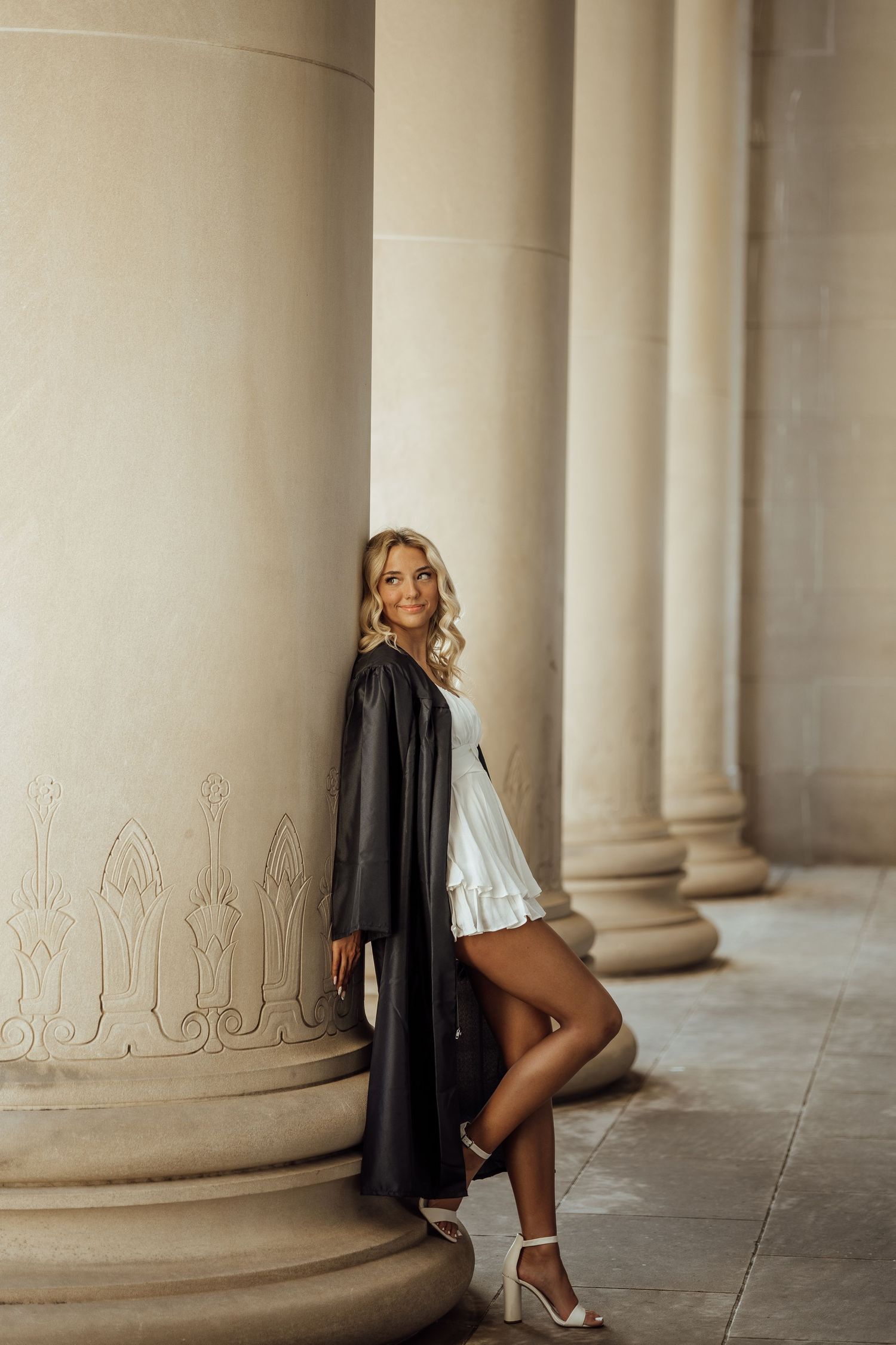 Blonde woman in white dress and black graduation gown poses beside ornate stone columns.