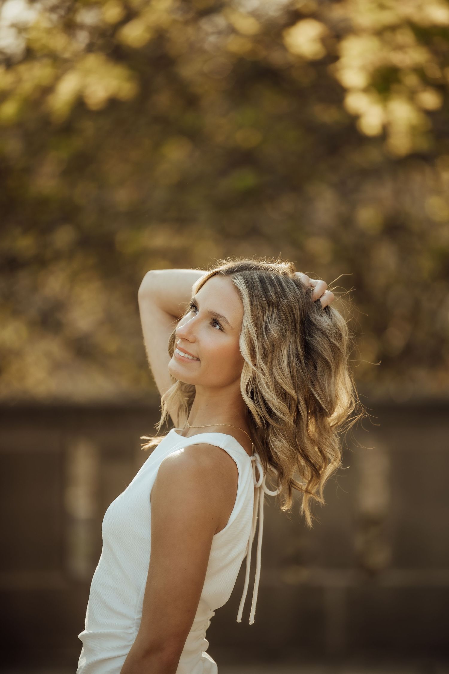 Smiling young woman with wavy blonde hair in white dress poses outdoors in golden hour light.