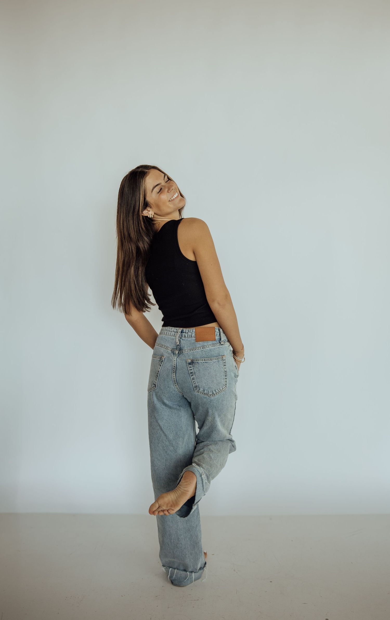 Young woman in black tank top and light wash jeans smiling while leaning against white wall.