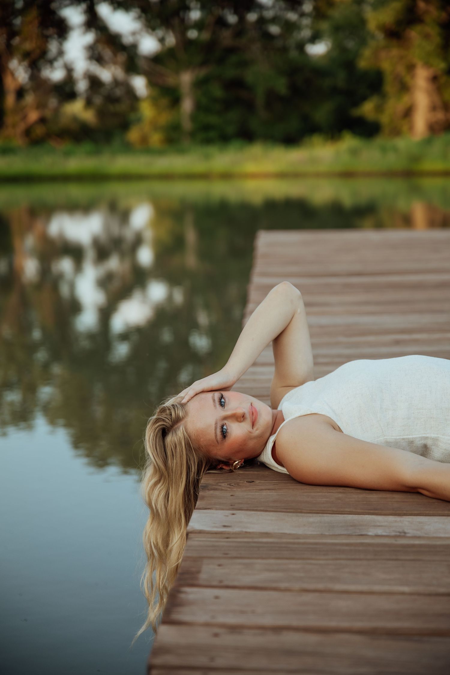 Blonde woman in white dress lying on wooden dock by calm reflective lake surrounded by green trees.