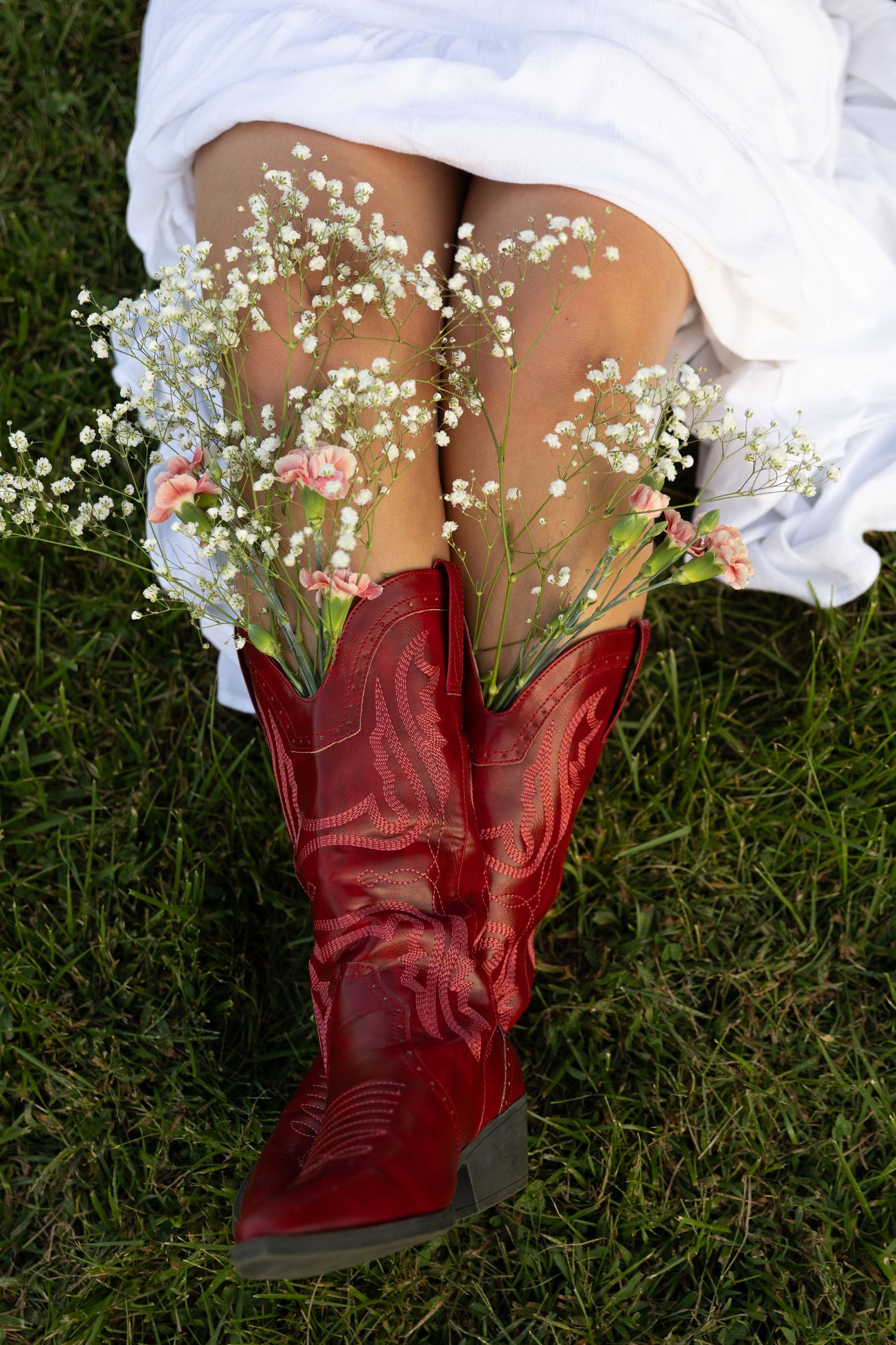 Red cowboy boots filled with baby's breath and pink flowers worn with a white dress on green grass.