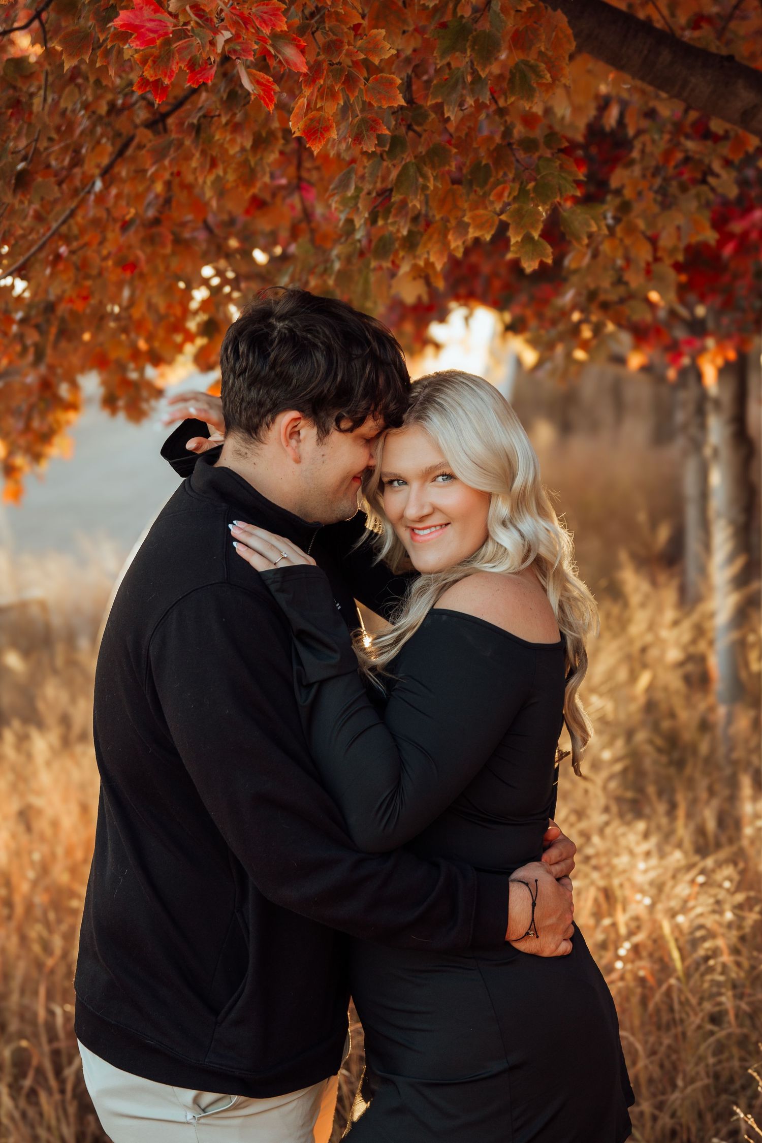 A couple embraces tenderly under vibrant red autumn leaves in a warm golden outdoor engagement portrait.