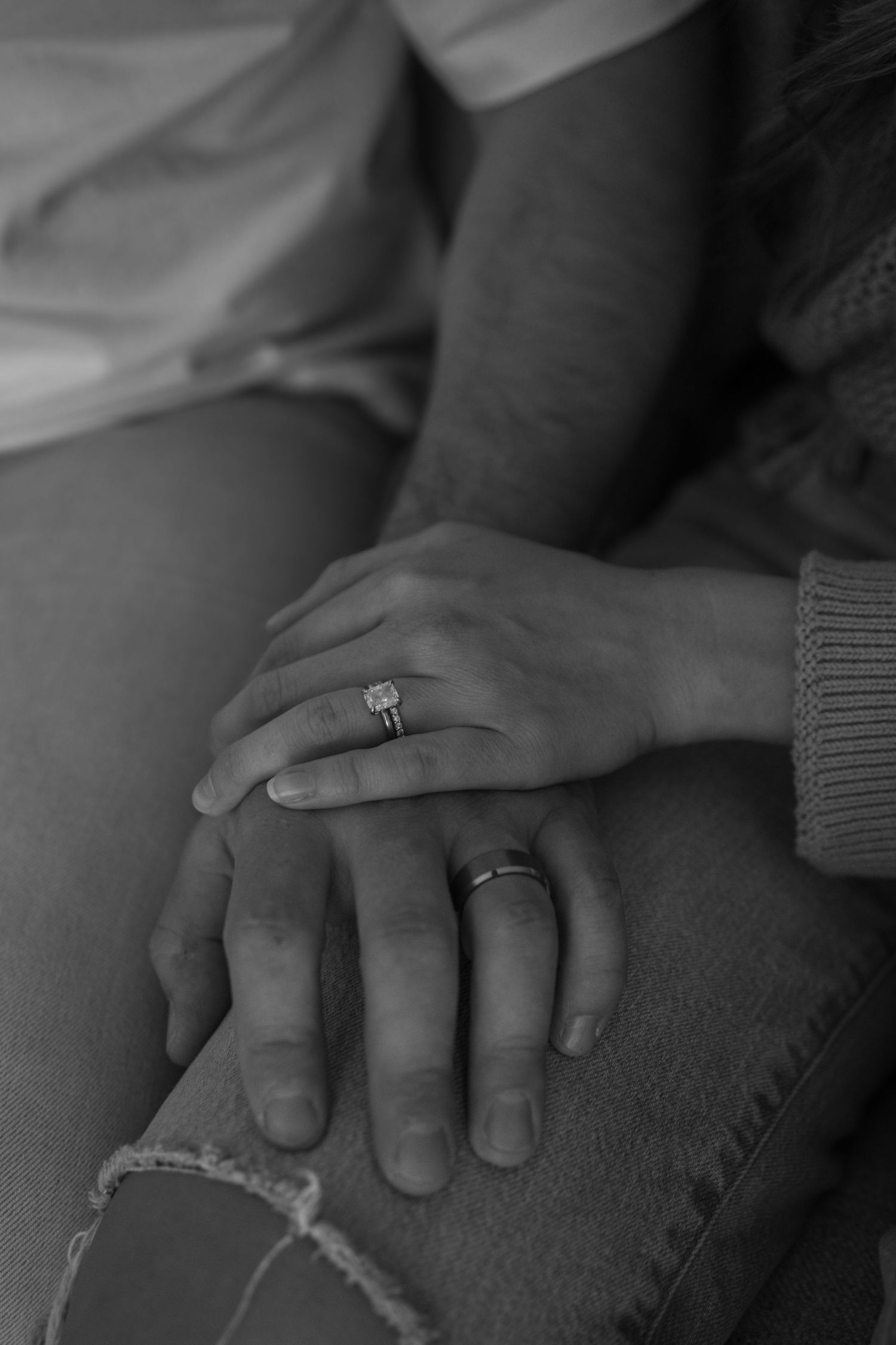 Black and white close-up of couple's hands showing engagement ring and wedding band resting on denim jeans.