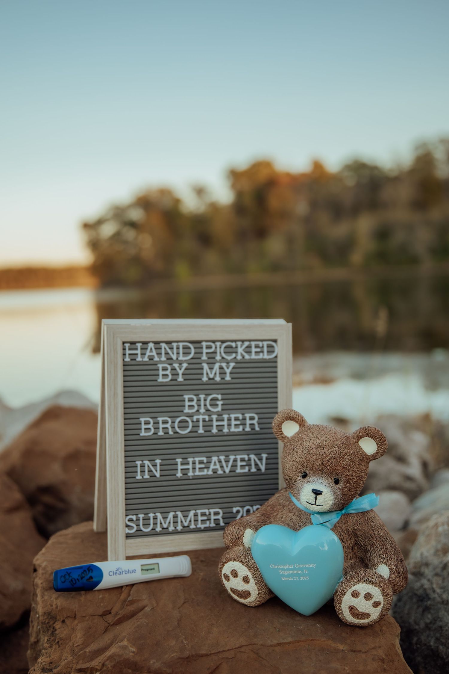 Pregnancy announcement sign reading Hand Picked By My Big Brother In Heaven with teddy bear by a lake.