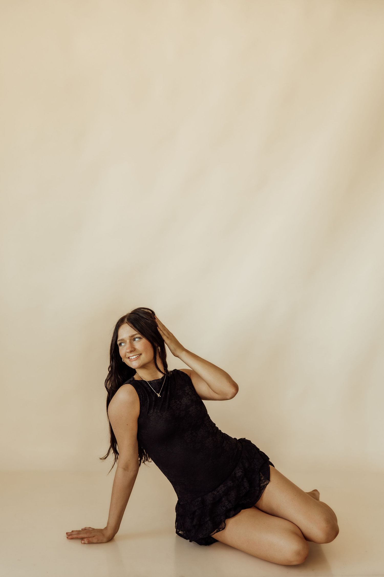 Young woman in black dress posing on floor against beige background, smiling with hand in hair.