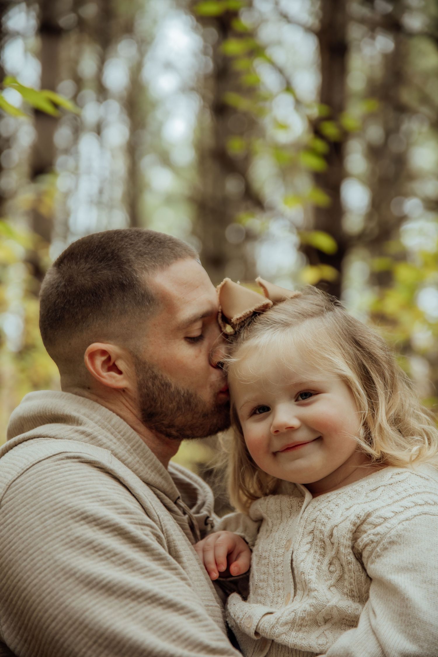 Father kissing his smiling toddler daughter on the forehead in a sunlit forest setting.