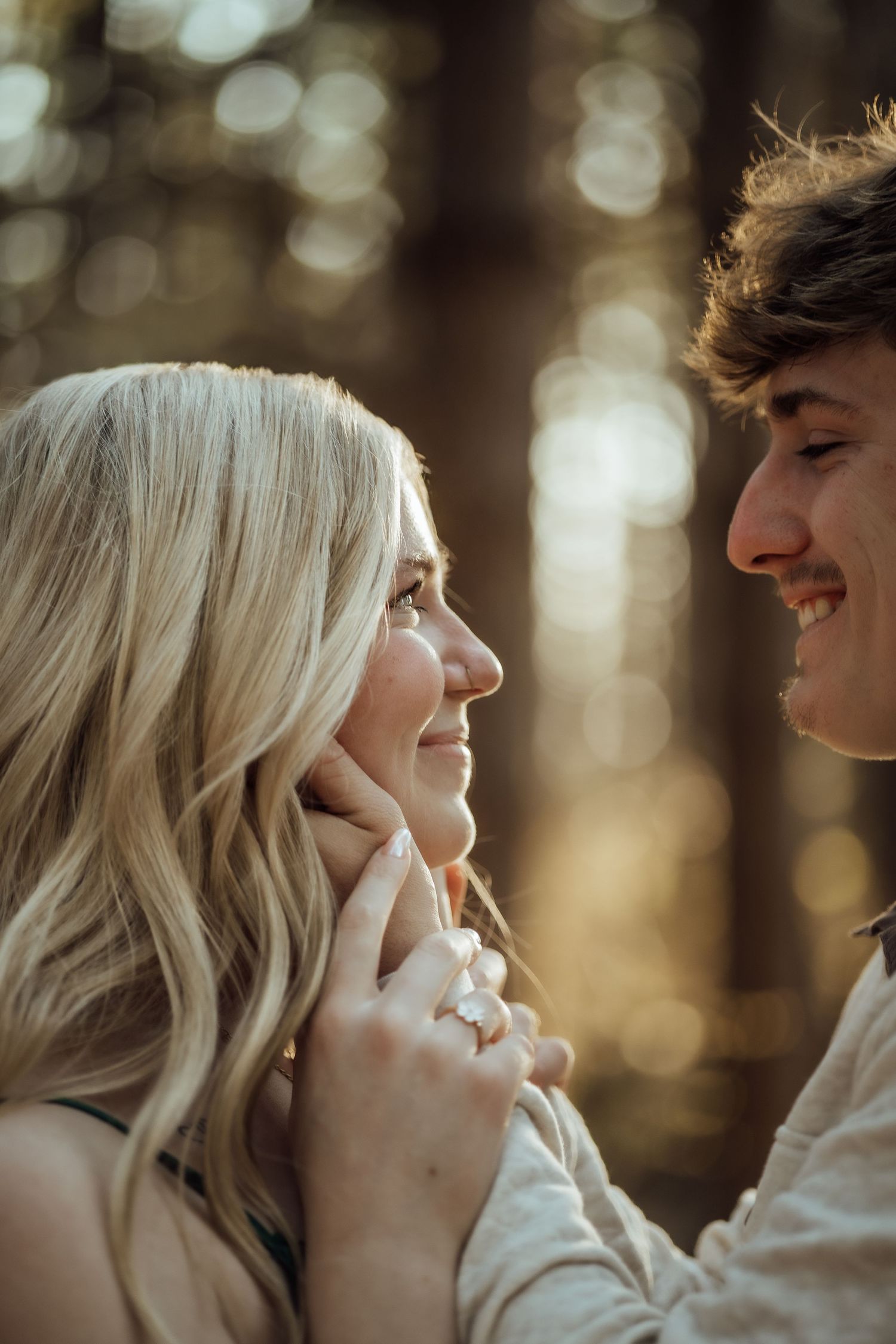 Smiling couple gazing at each other in a sun-drenched forest with warm bokeh background lighting.