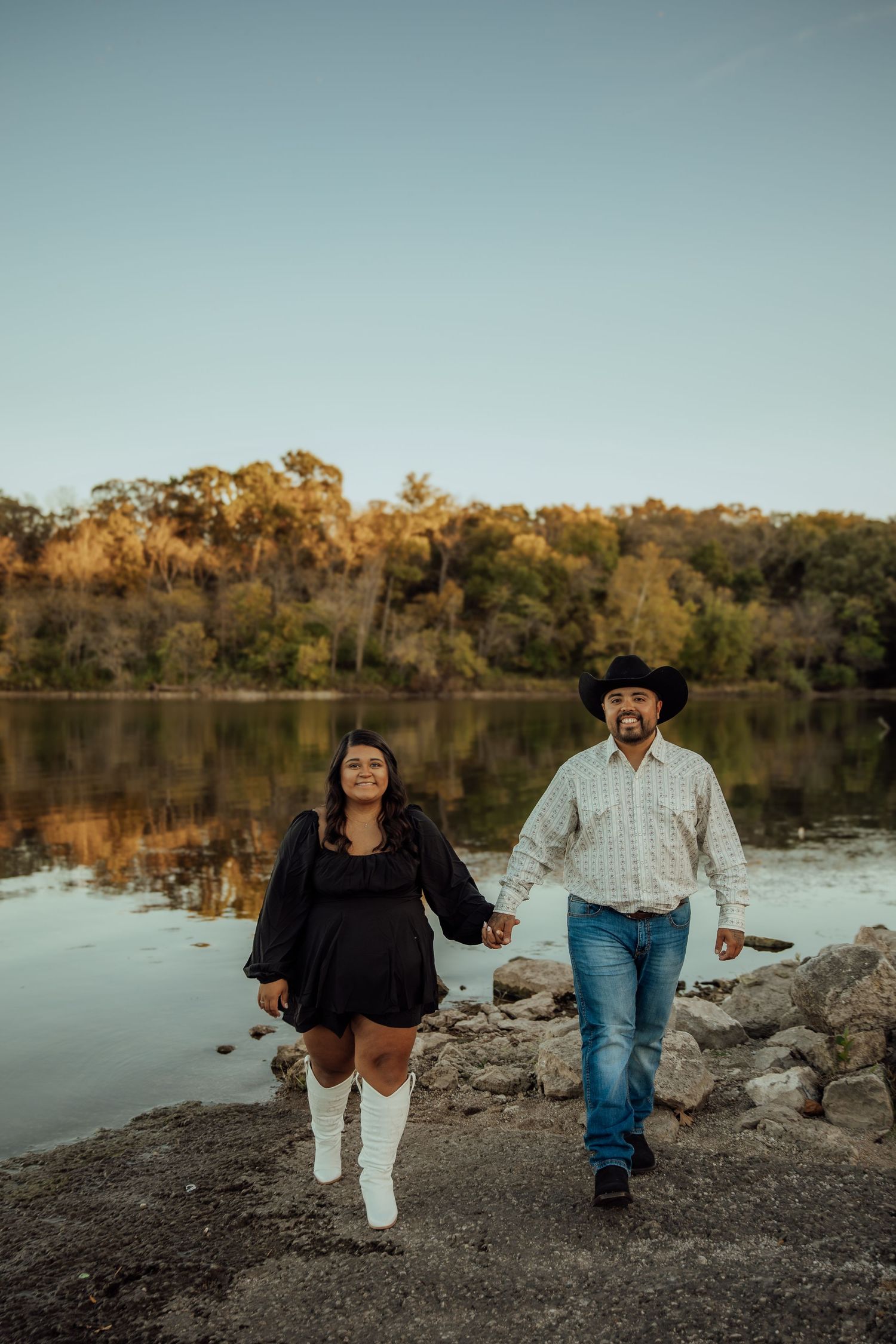 Couple holding hands walking along rocky lakeside at sunset, woman in black dress, man in cowboy hat.