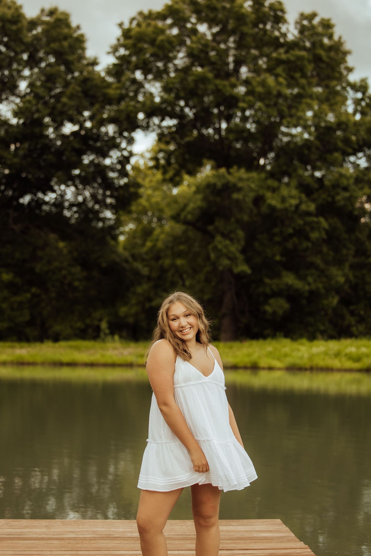 Young woman in white sundress smiling on wooden dock by serene lake with lush green trees backdrop.