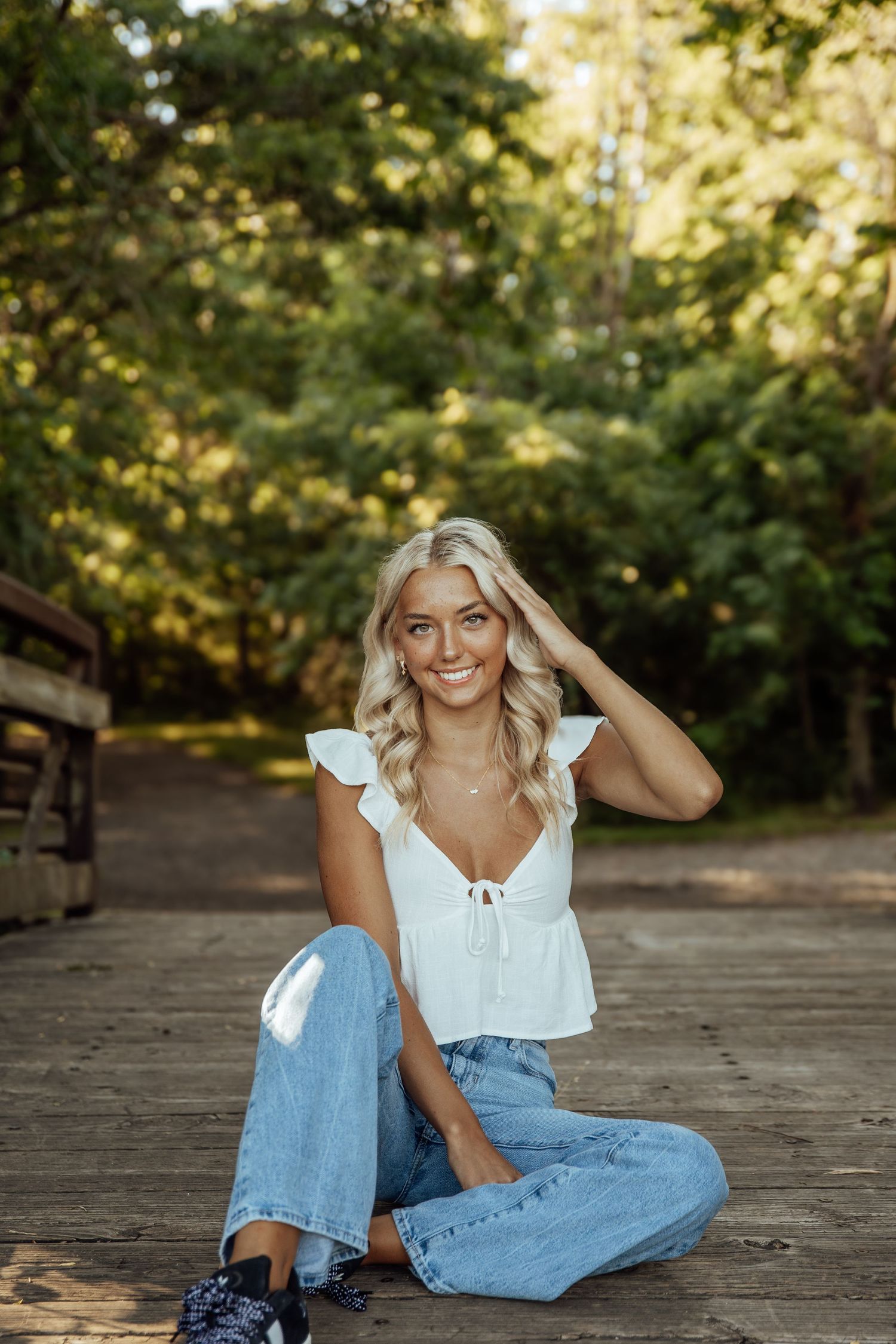 Smiling blonde woman in white top and jeans sitting cross-legged on wooden bridge in sunny forest.