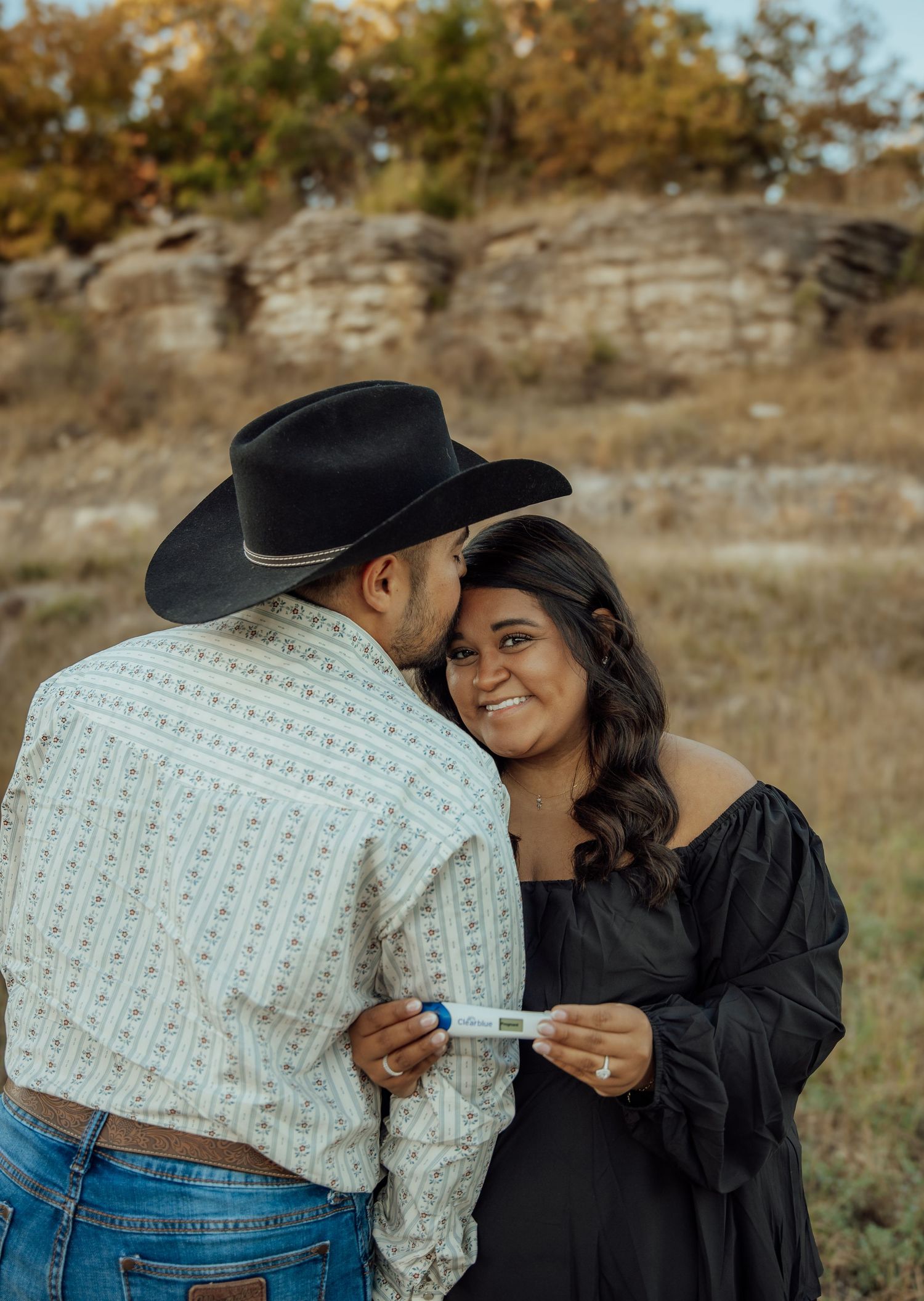 Couple sharing a joyful pregnancy announcement moment outdoors, man in cowboy hat kissing womans cheek.
