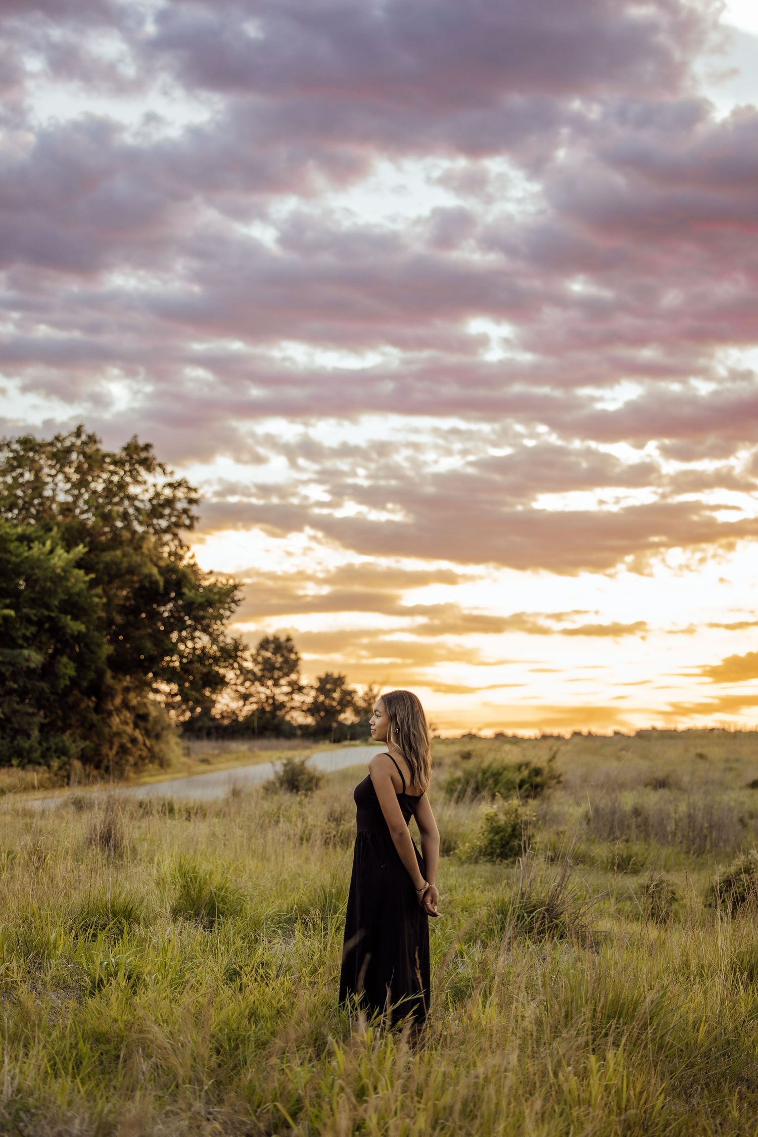 Woman in black dress standing in grassy field at golden sunset with dramatic cloudy sky.