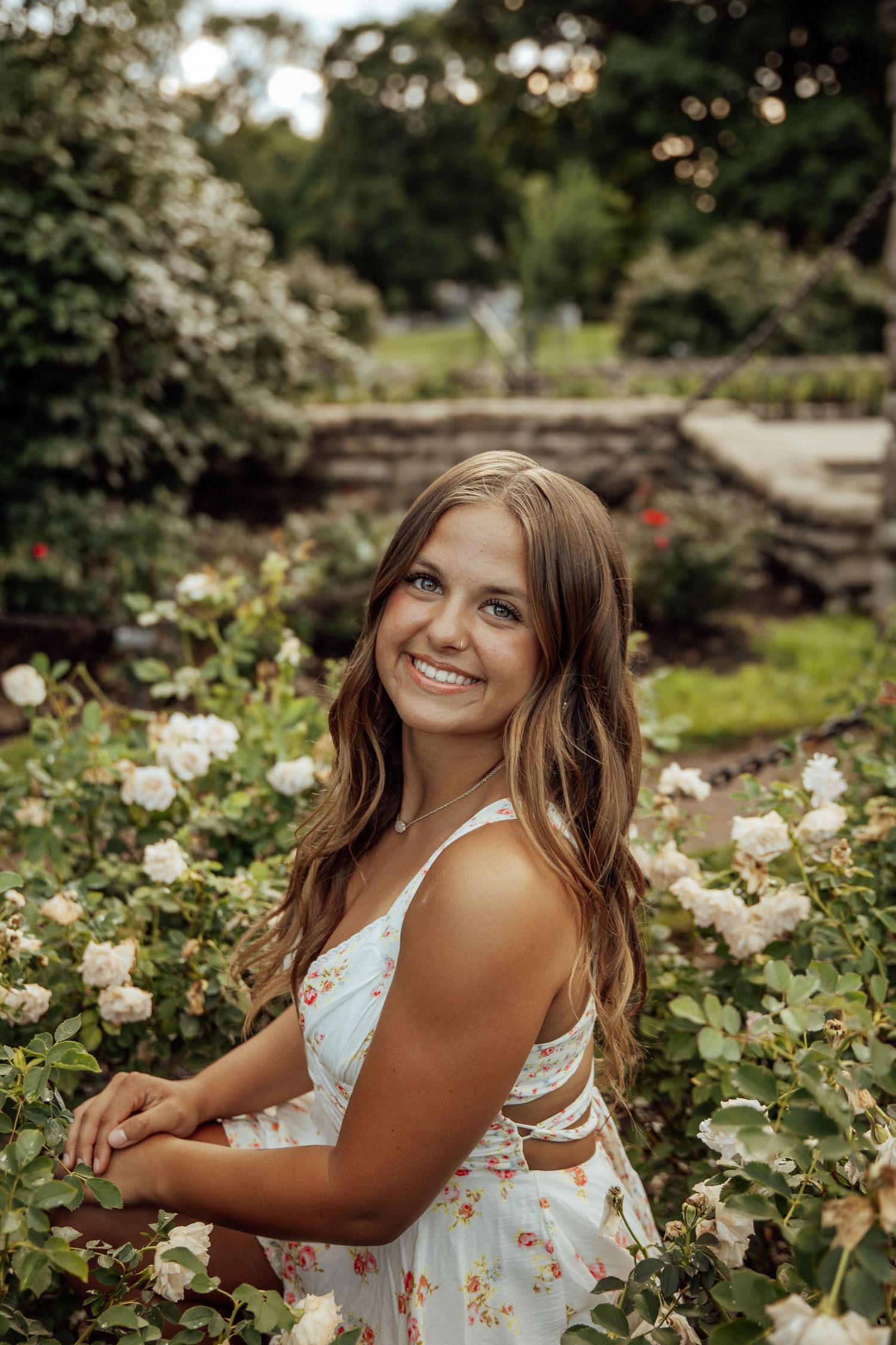 Smiling brunette woman in floral sundress posing among white rose bushes in a lush garden.