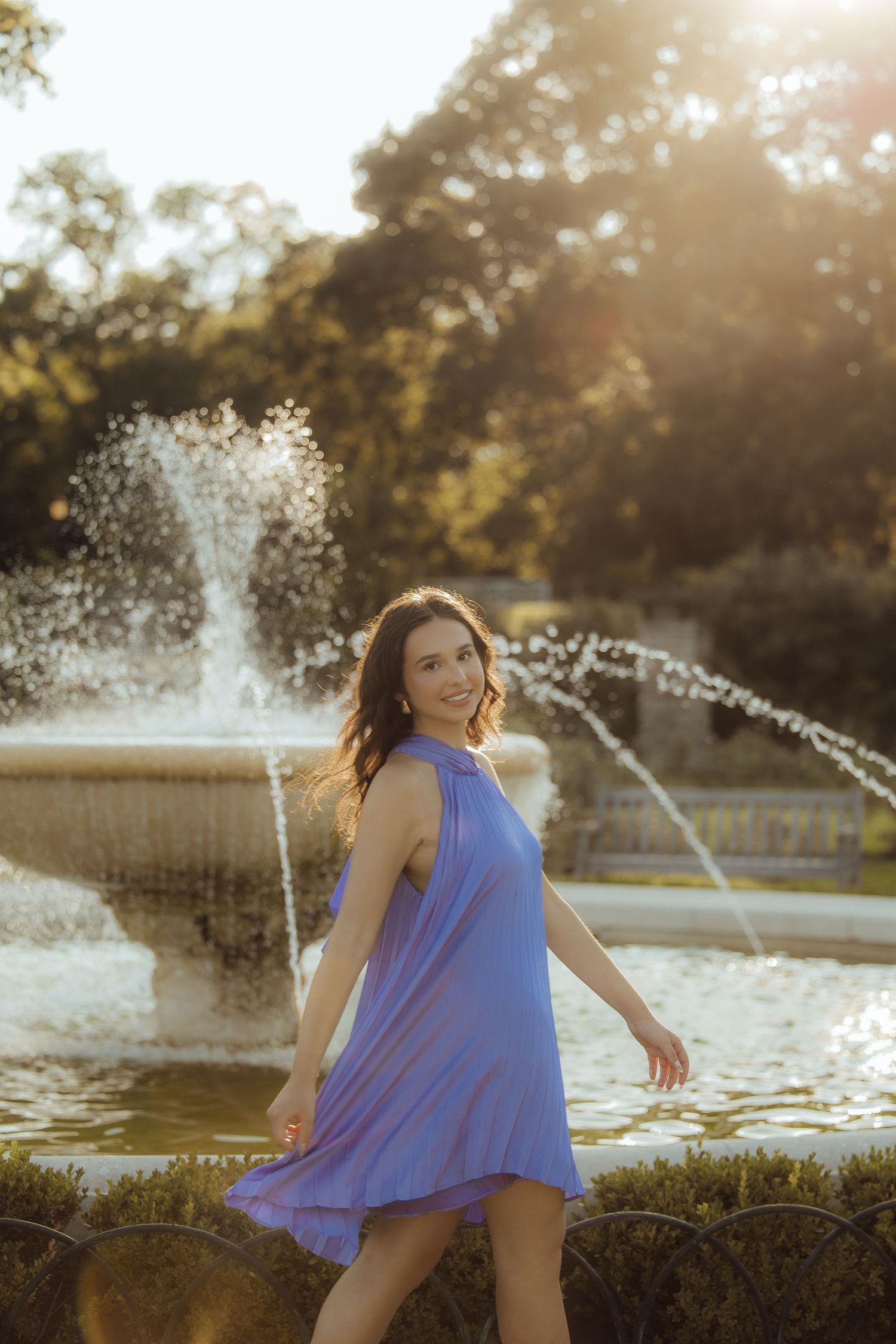 Woman in blue dress twirling by a sunlit fountain in a park.