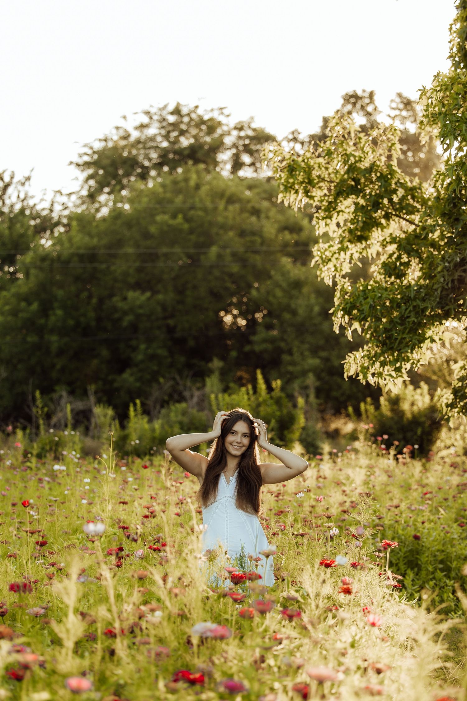 Young woman in light blue dress smiling in a vibrant wildflower meadow with lush green trees at golden hour.