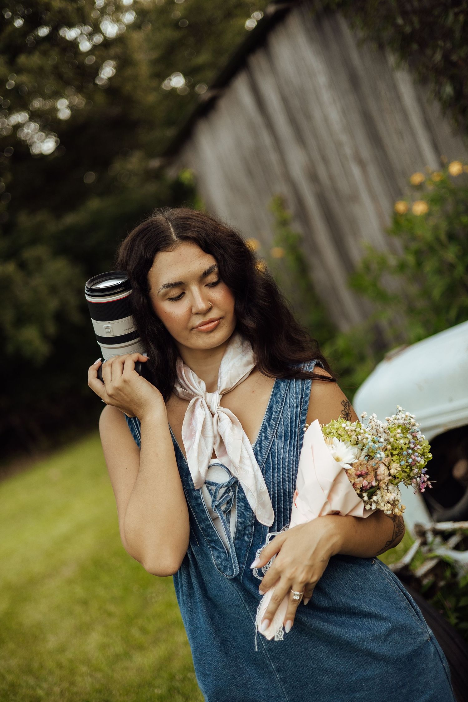 Woman in denim overalls holding a camera lens and wildflower bouquet outdoors near a vintage car.
