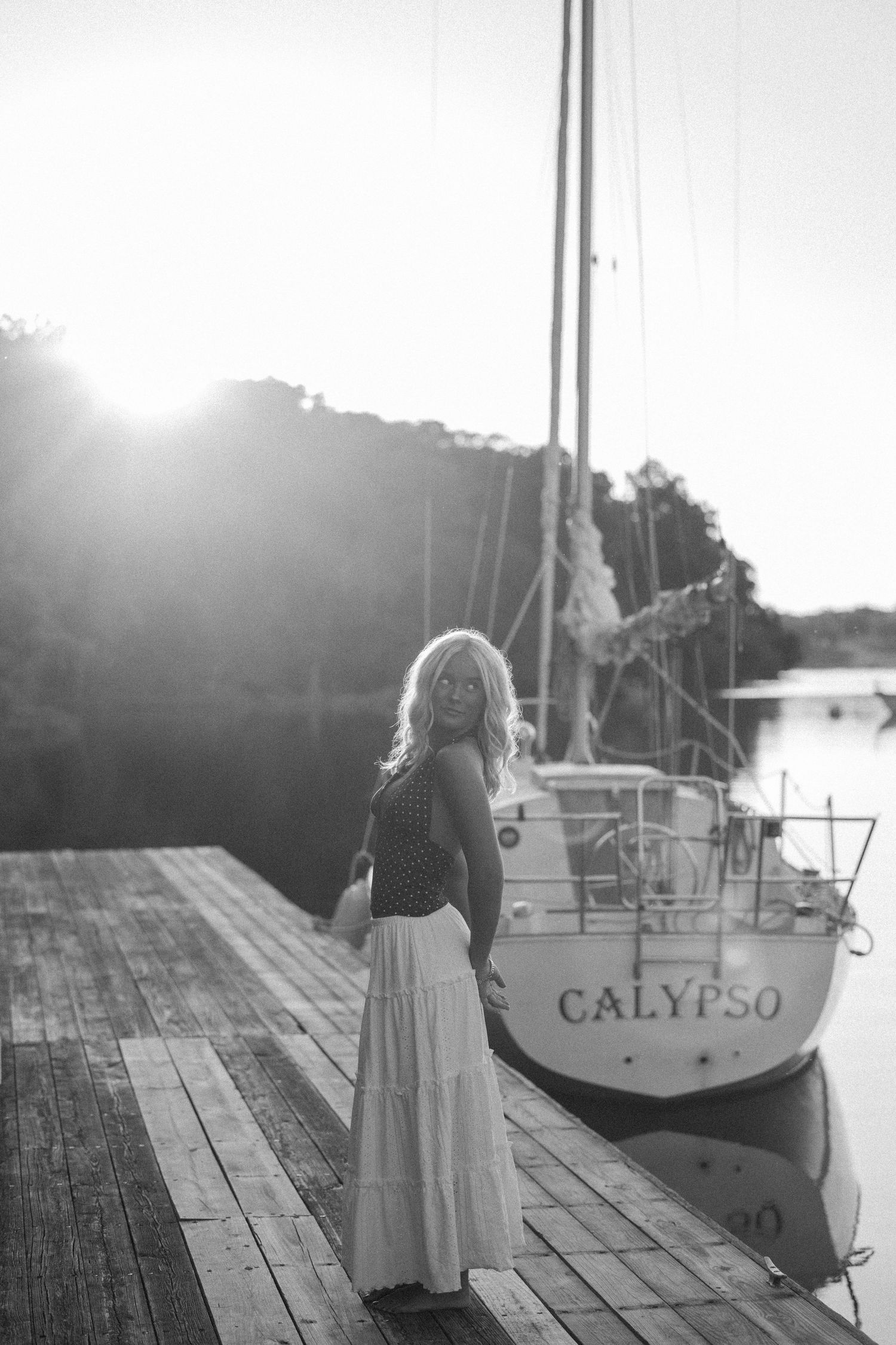 Black and white photo of a woman in a maxi skirt standing on a dock beside a sailboat named Calypso at sunset.