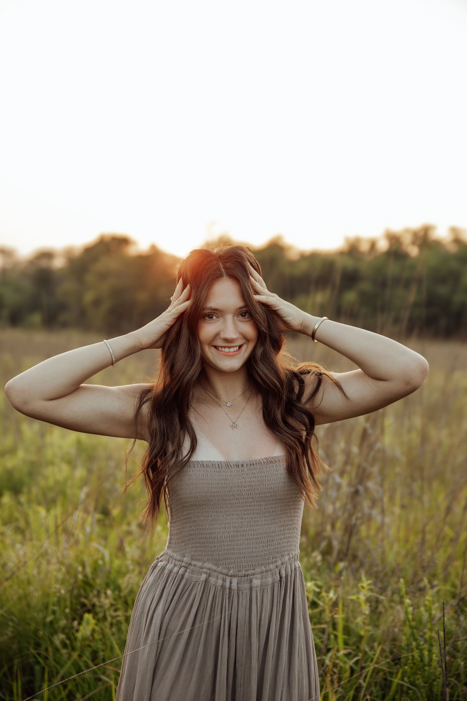 Smiling brunette woman in gray smocked dress standing in golden field at sunset with hands in hair.