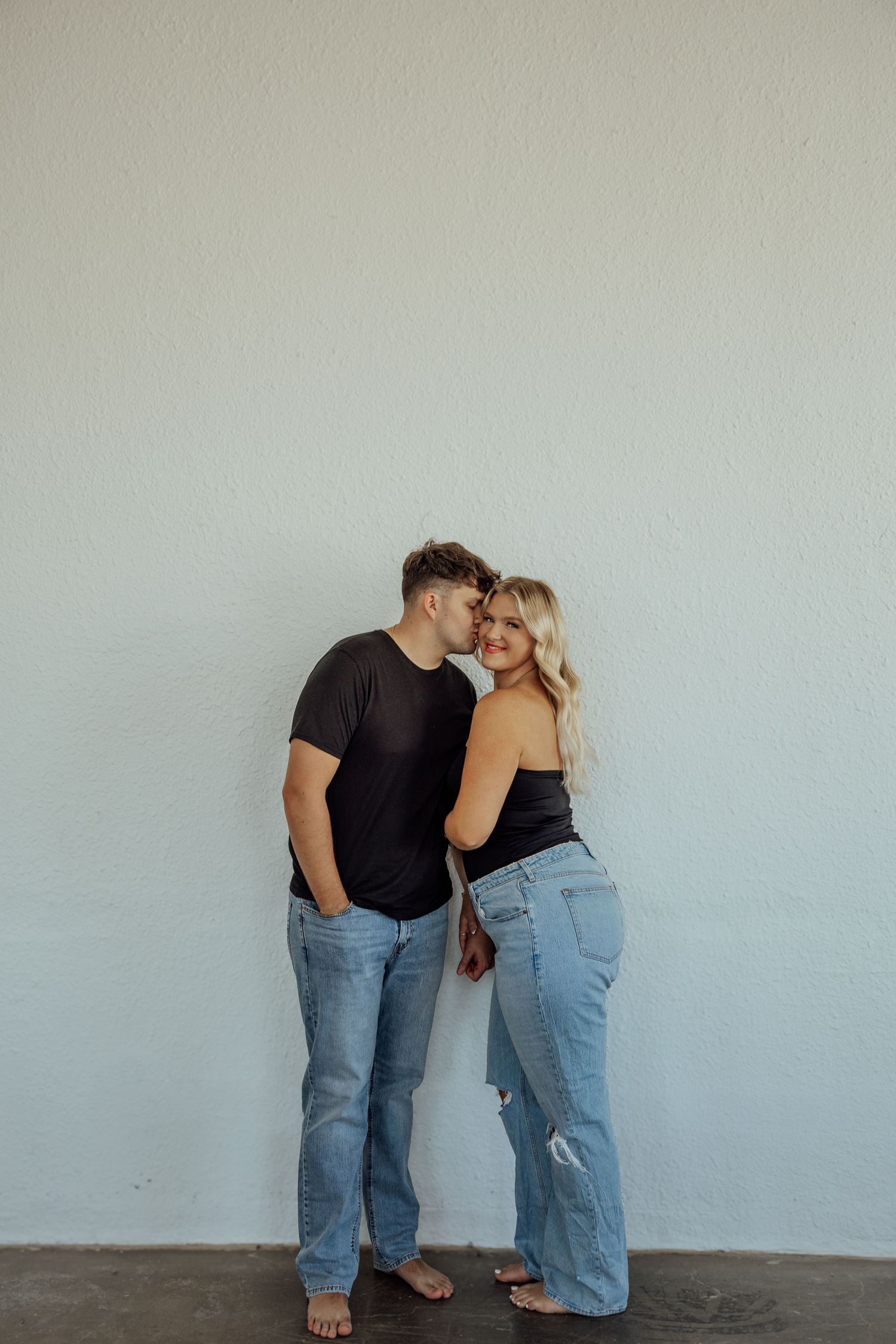Couple leaning against white wall, man in black tee kissing woman's temple, both wearing jeans.