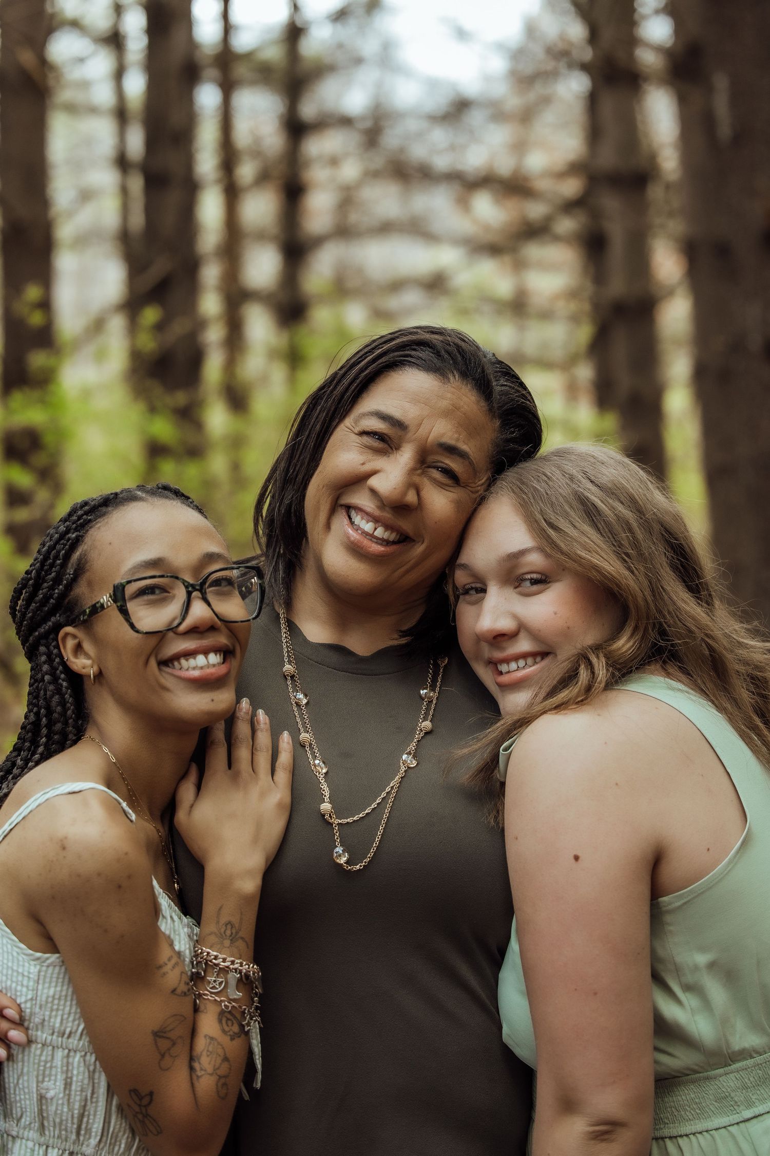Three women smiling and embracing each other during an outdoor family portrait session in the woods.