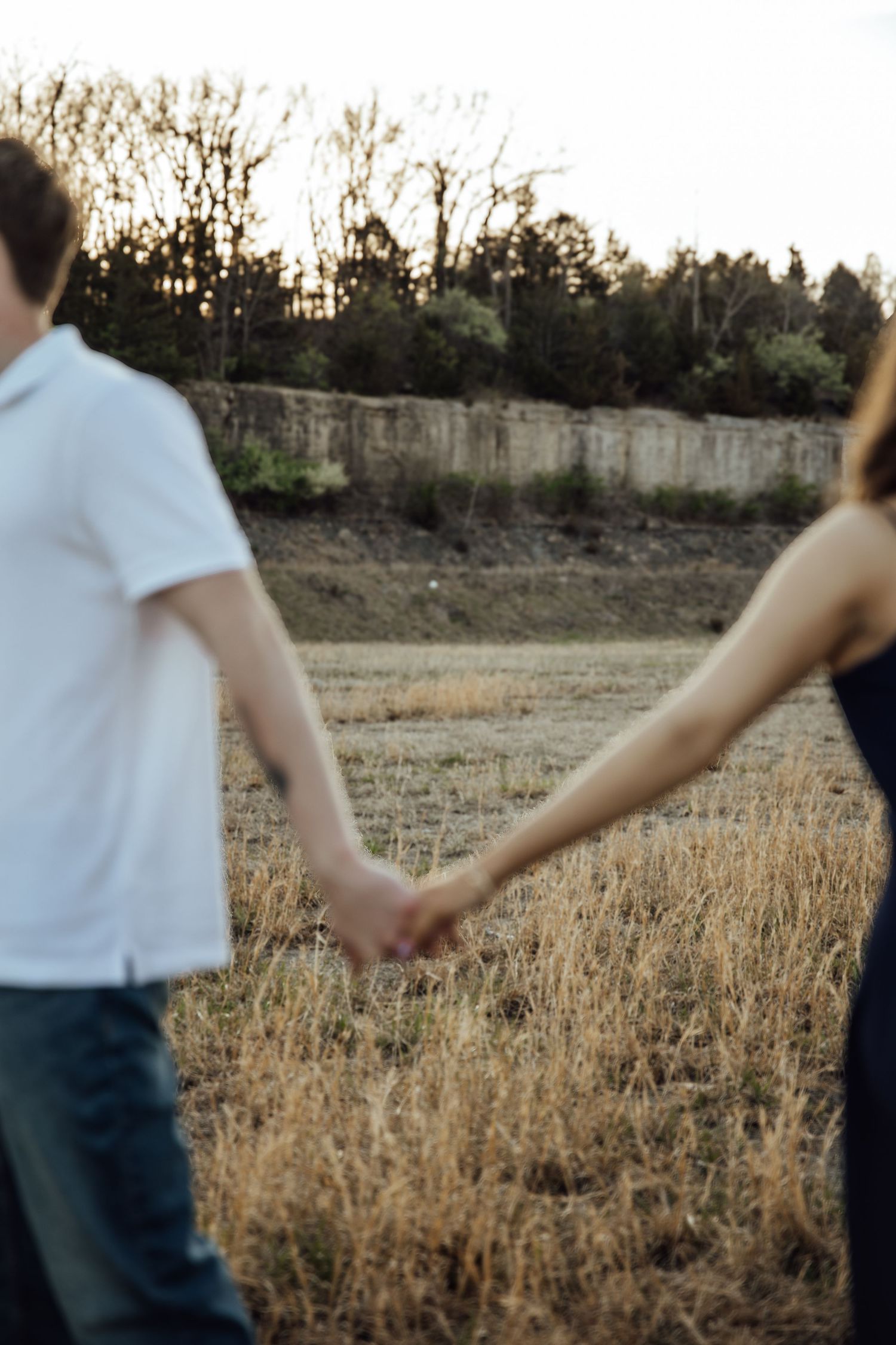 A couple holds hands while walking through a dry golden field at sunset during an outdoor engagement photo session.