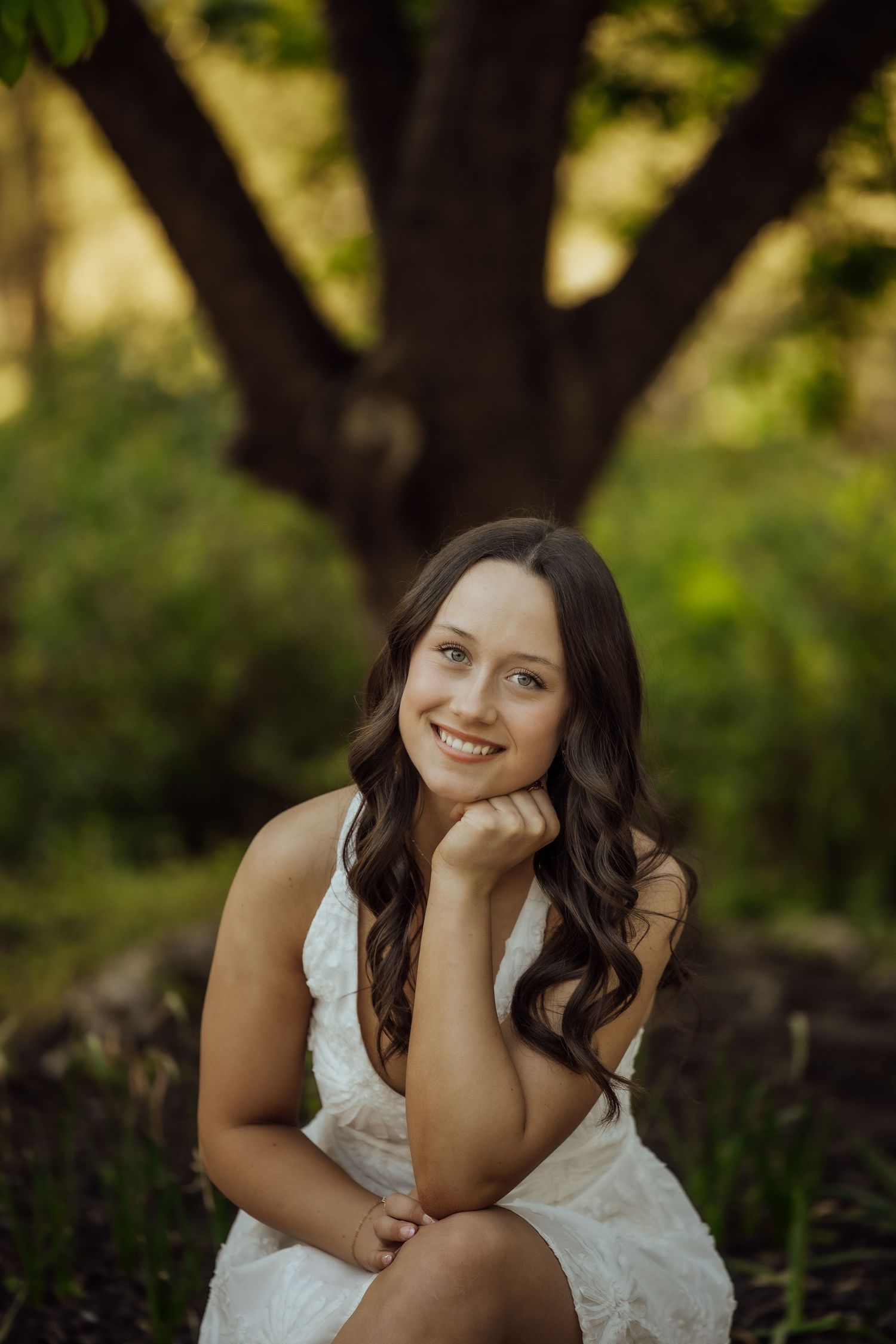 Brunette woman in white dress smiles warmly while seated outdoors near a large tree.