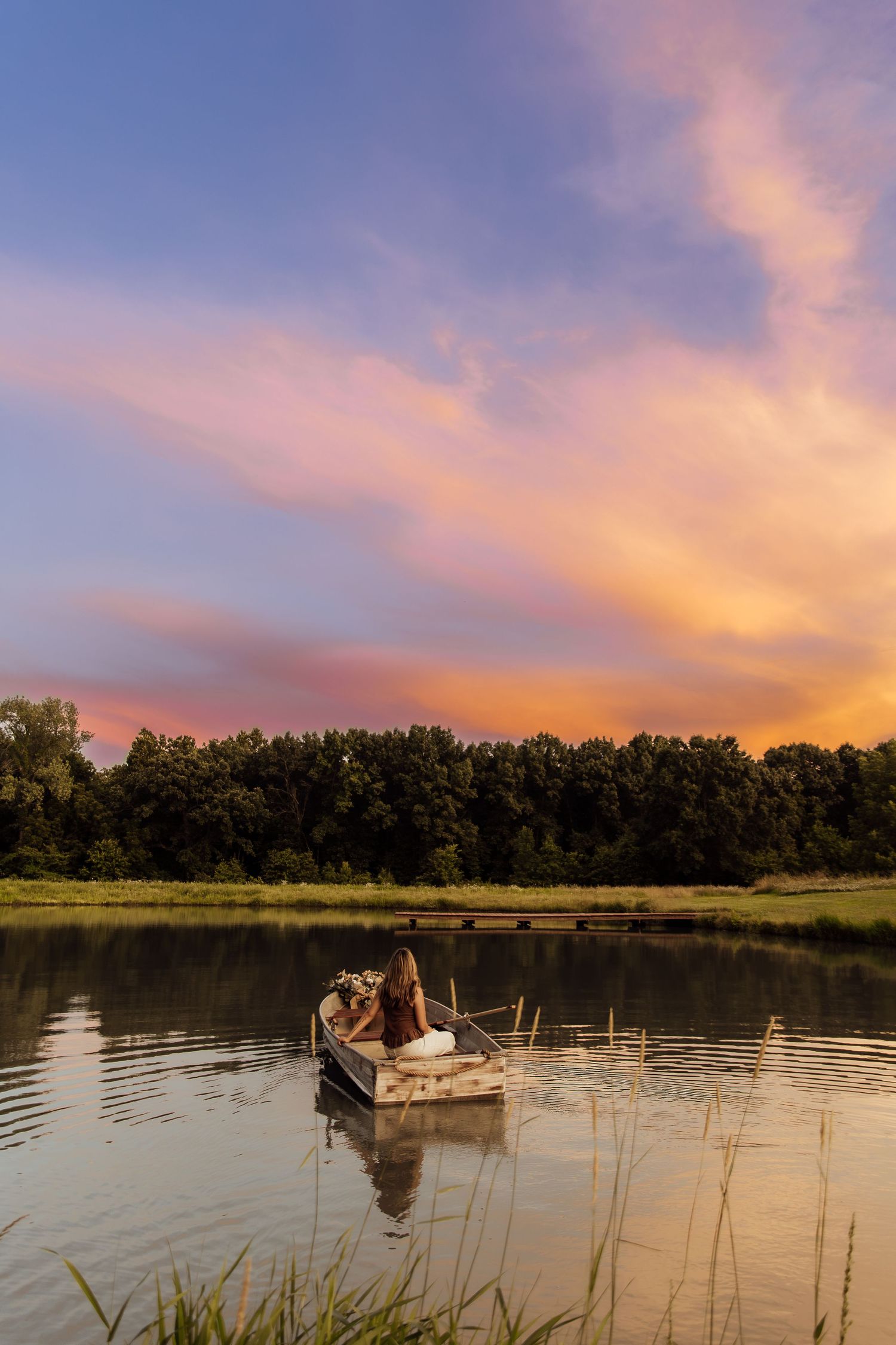 Couple sitting in a rowboat on a calm lake at sunset with vibrant pink and orange clouds reflected in water.