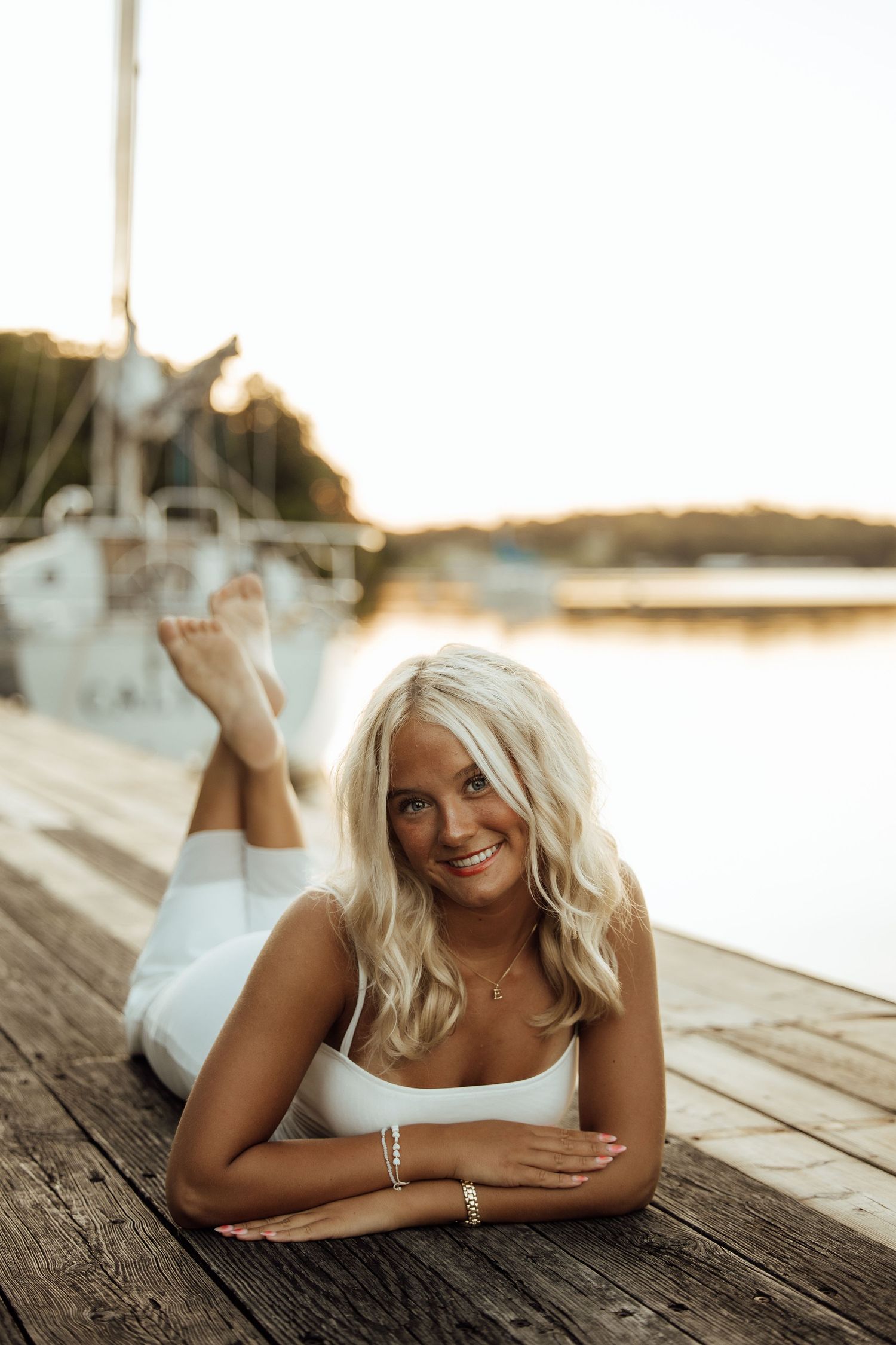 Smiling blonde woman in white outfit lying on a wooden dock at sunset with sailboats in the background.