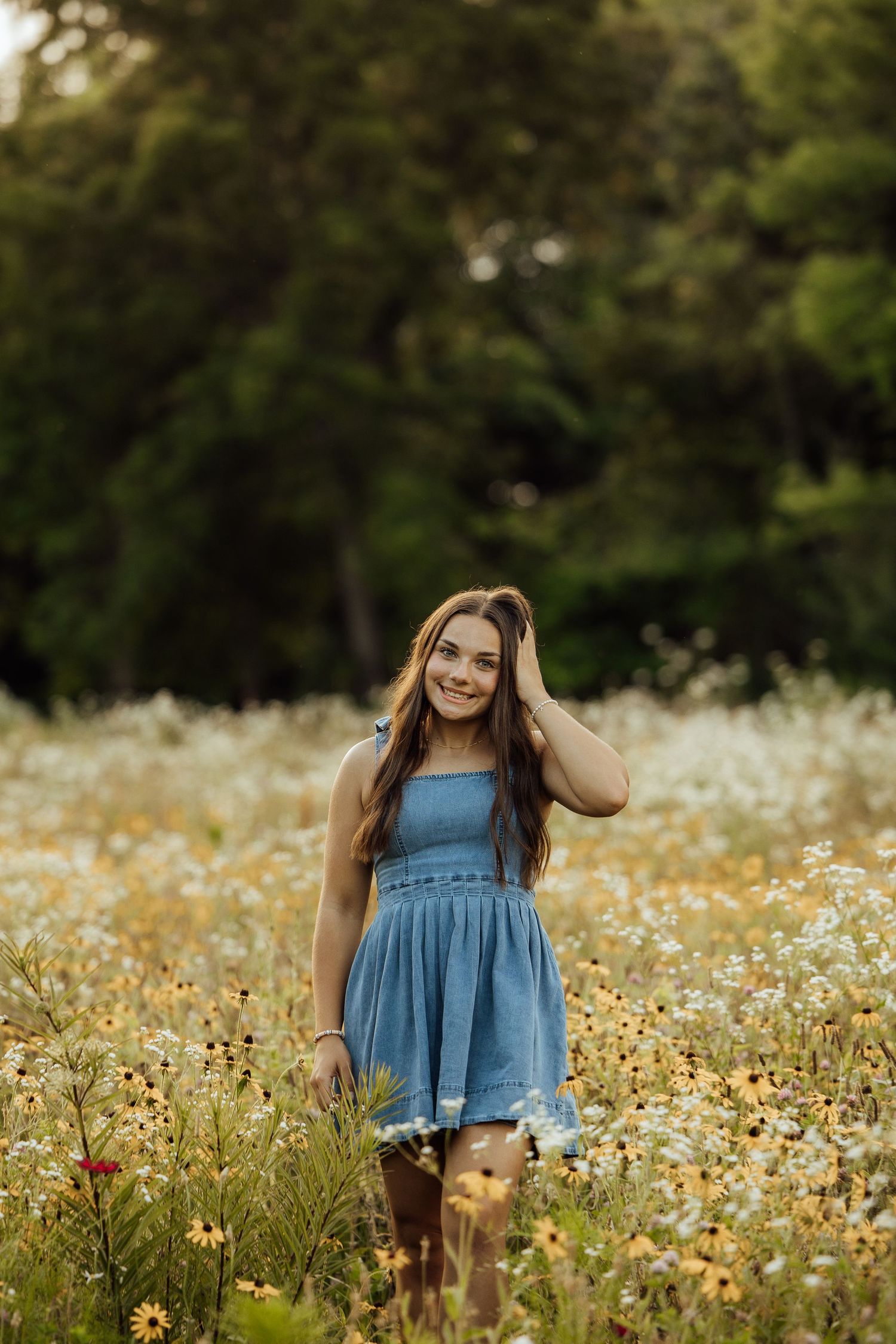 Young woman in blue dress smiling in a wildflower field with yellow and white blooms and green trees.