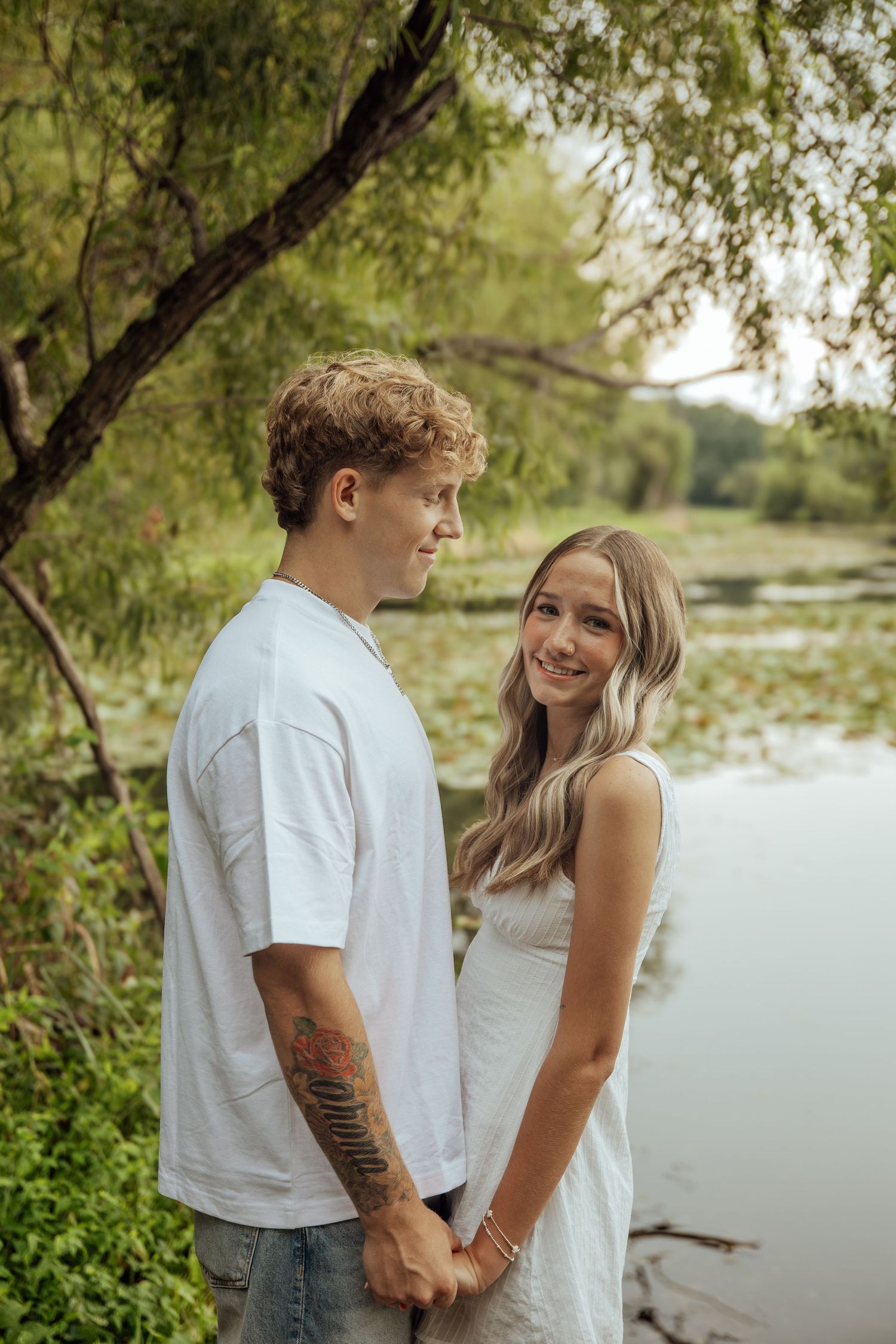 A young couple holds hands near a serene pond surrounded by lush green trees and foliage.