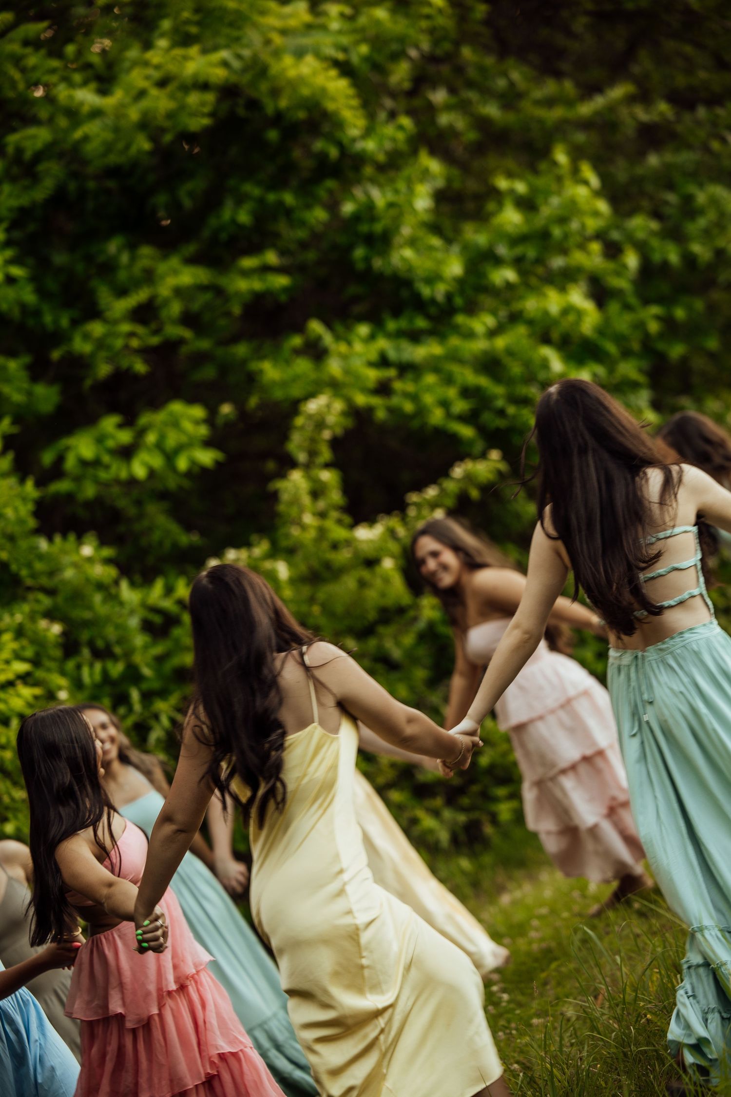 Young women in pastel dresses dancing and holding hands in a lush green outdoor setting.