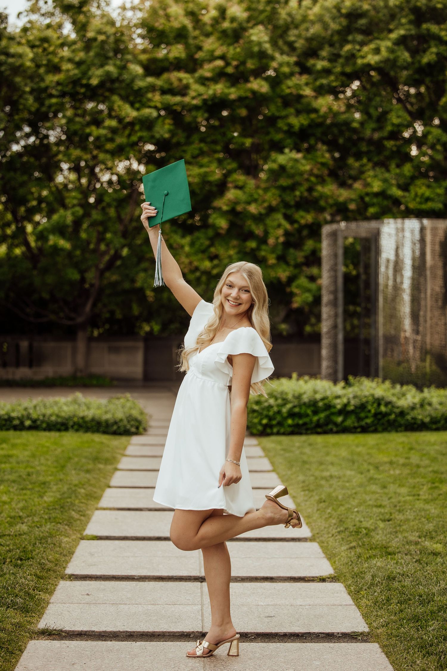 Smiling blonde graduate in white dress holds green cap overhead on garden pathway.
