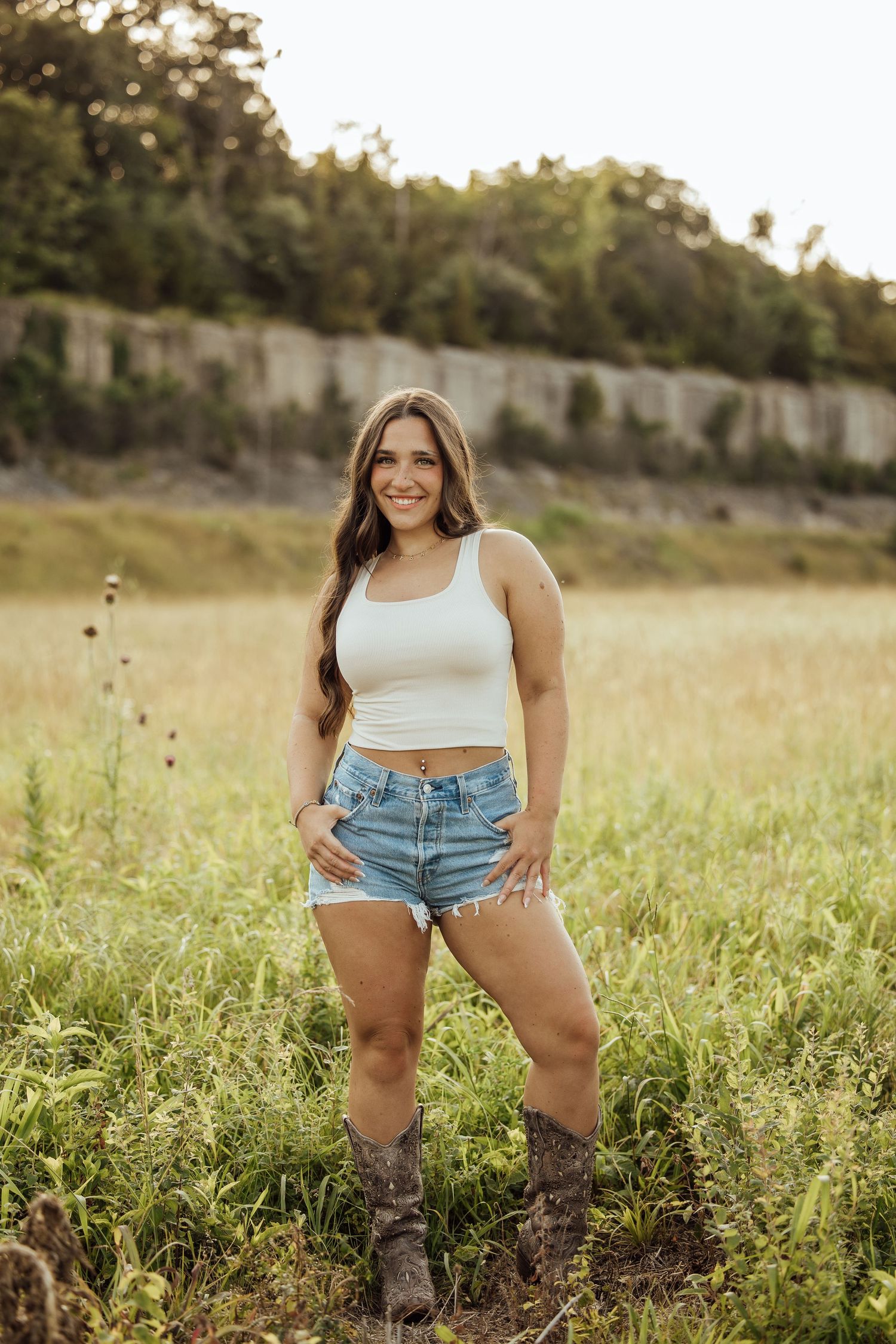 Young woman in white crop top, denim shorts, and cowboy boots posing in a sunlit green field with rocky cliffs behind her.