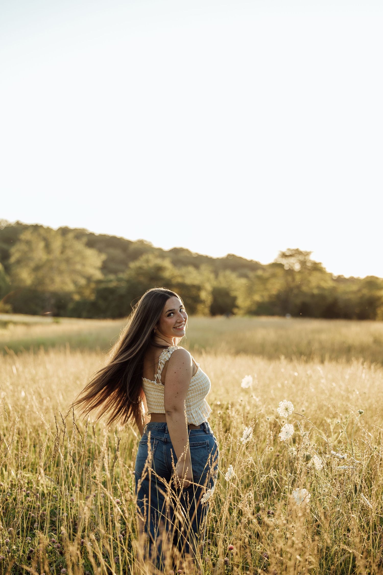 Young woman with long hair in white crop top and jeans smiling in golden sunlit field at sunset.