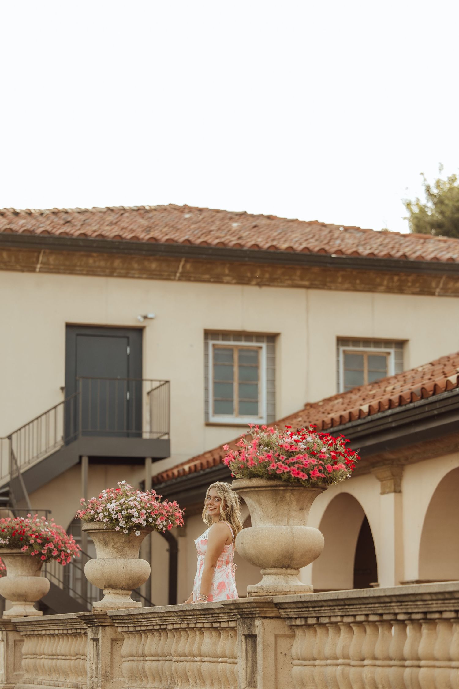 Woman in pink dress standing among large stone urns with red flowers outside a Mediterranean-style building.