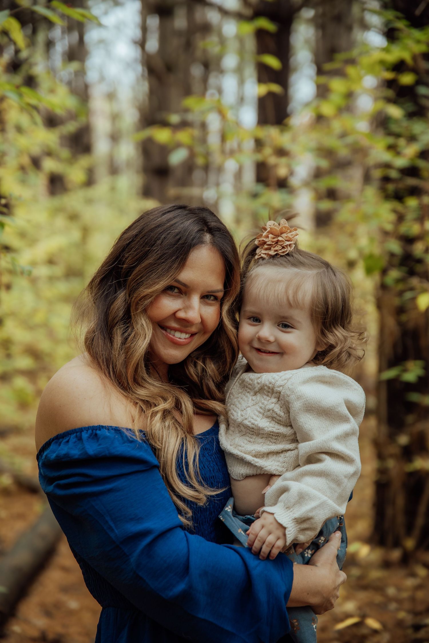 Smiling mother holding her young daughter with a bow in her hair in an autumn forest.