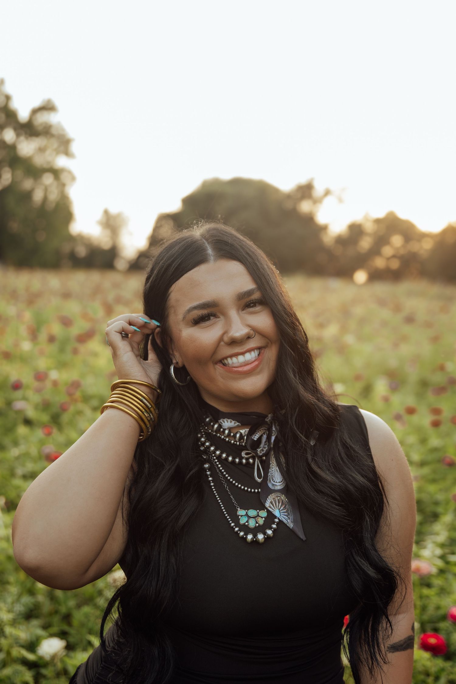 Smiling dark-haired woman in black dress with layered turquoise jewelry in a sunlit flower field.