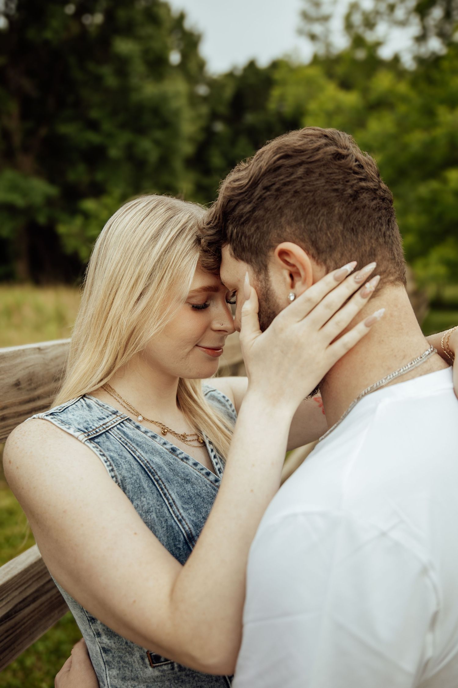 A romantic couple touches foreheads tenderly near a wooden fence in a lush green outdoor setting.