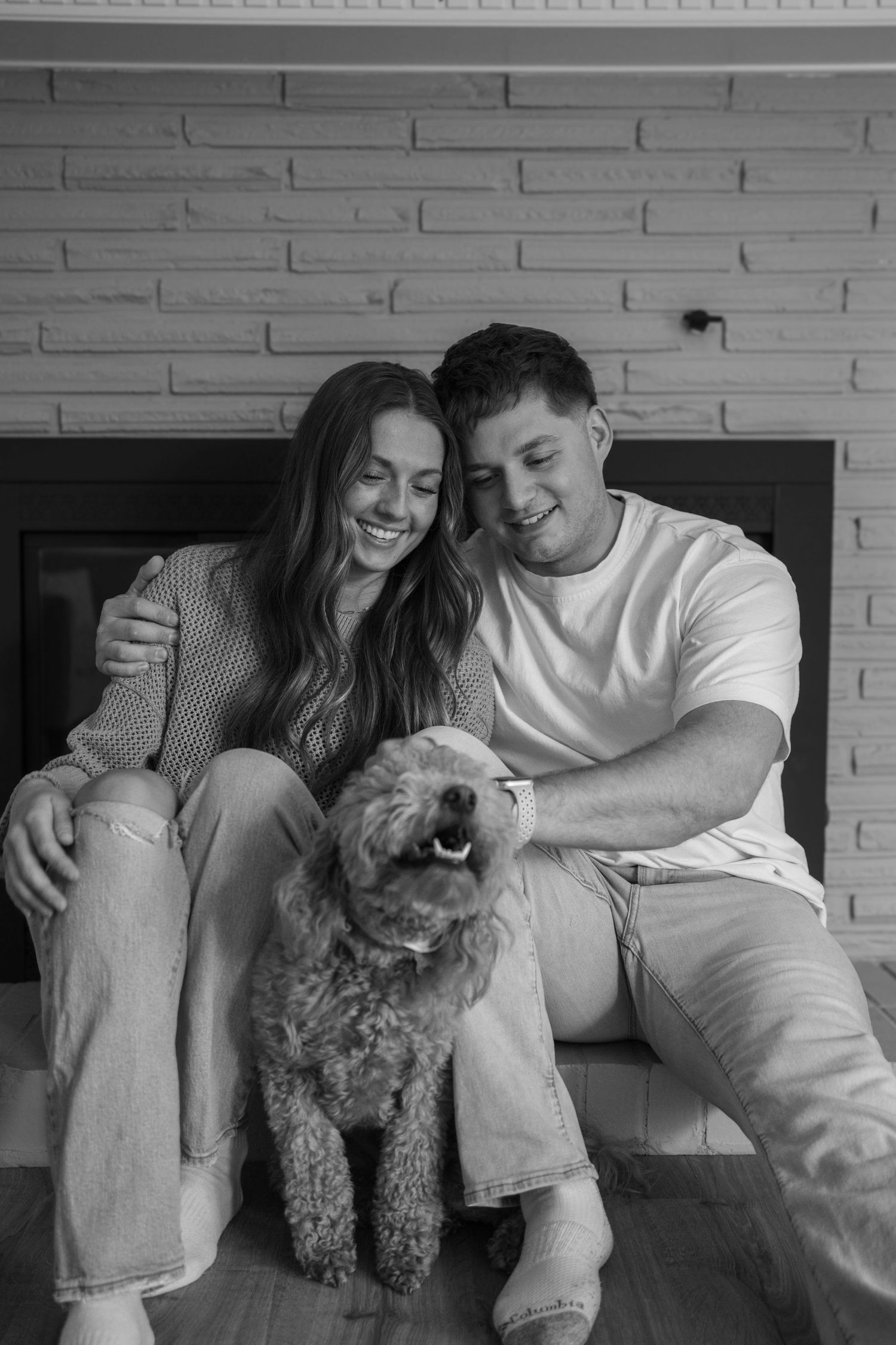 Black and white photo of smiling couple sitting by fireplace with their fluffy poodle dog.