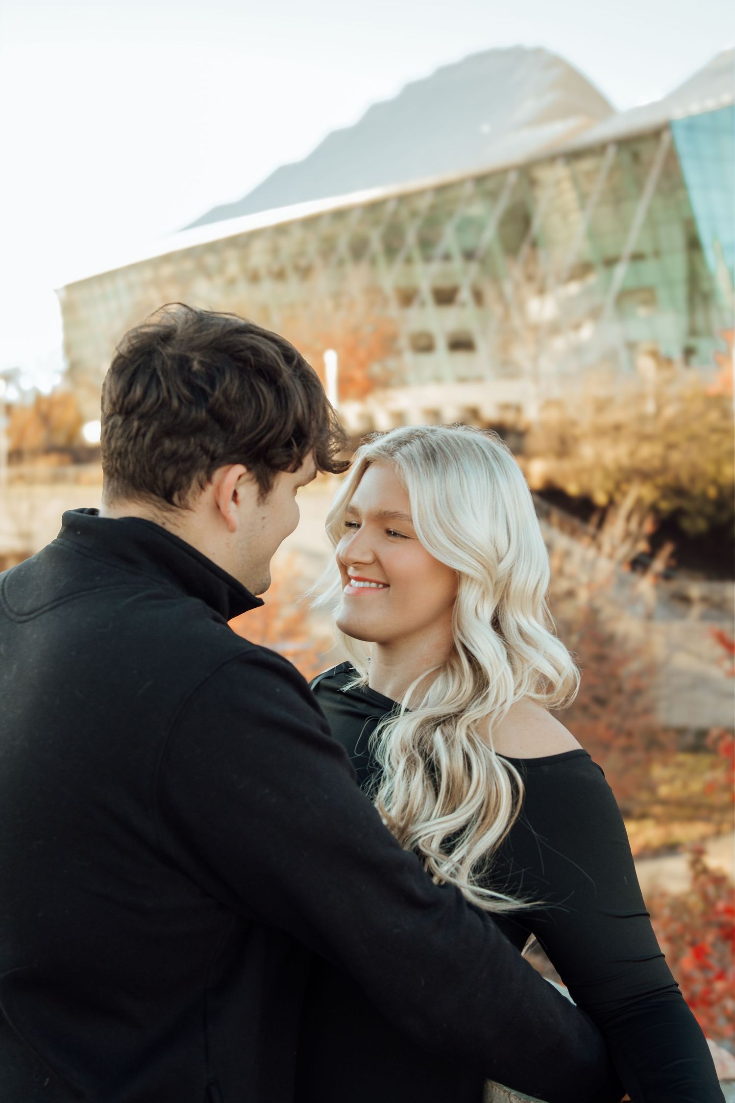 A smiling blonde woman in a black off-shoulder dress shares a close romantic moment with her partner outdoors.