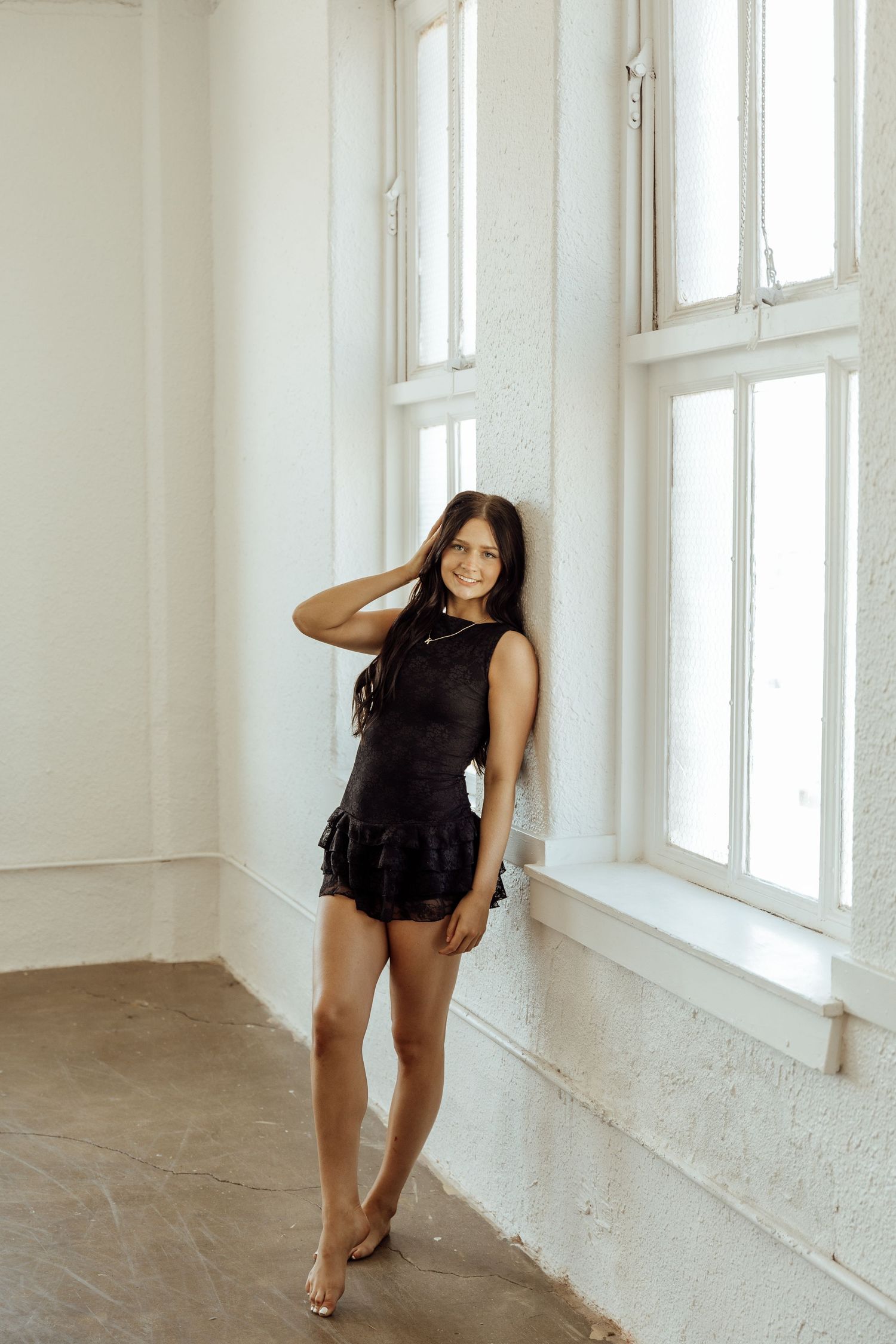 Young woman in black mini dress posing barefoot against white brick wall near large windows in bright studio.