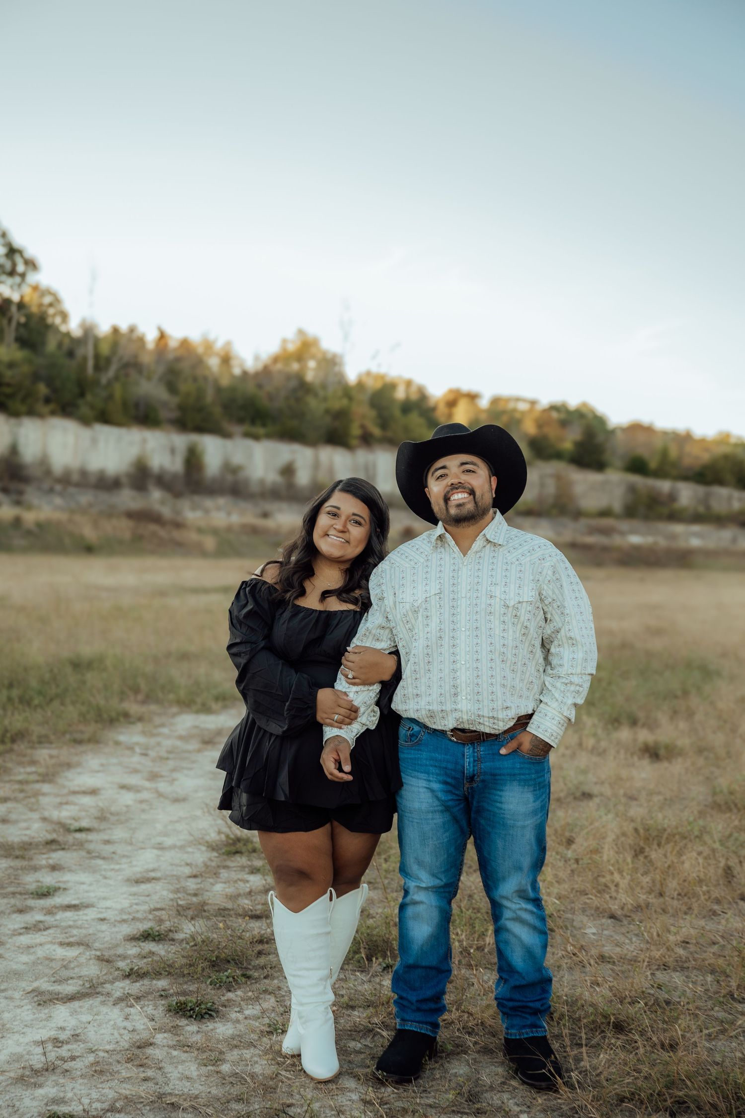 Couple posing in field at dusk, woman in black dress with white cowboy boots, man in cowboy hat and jeans.