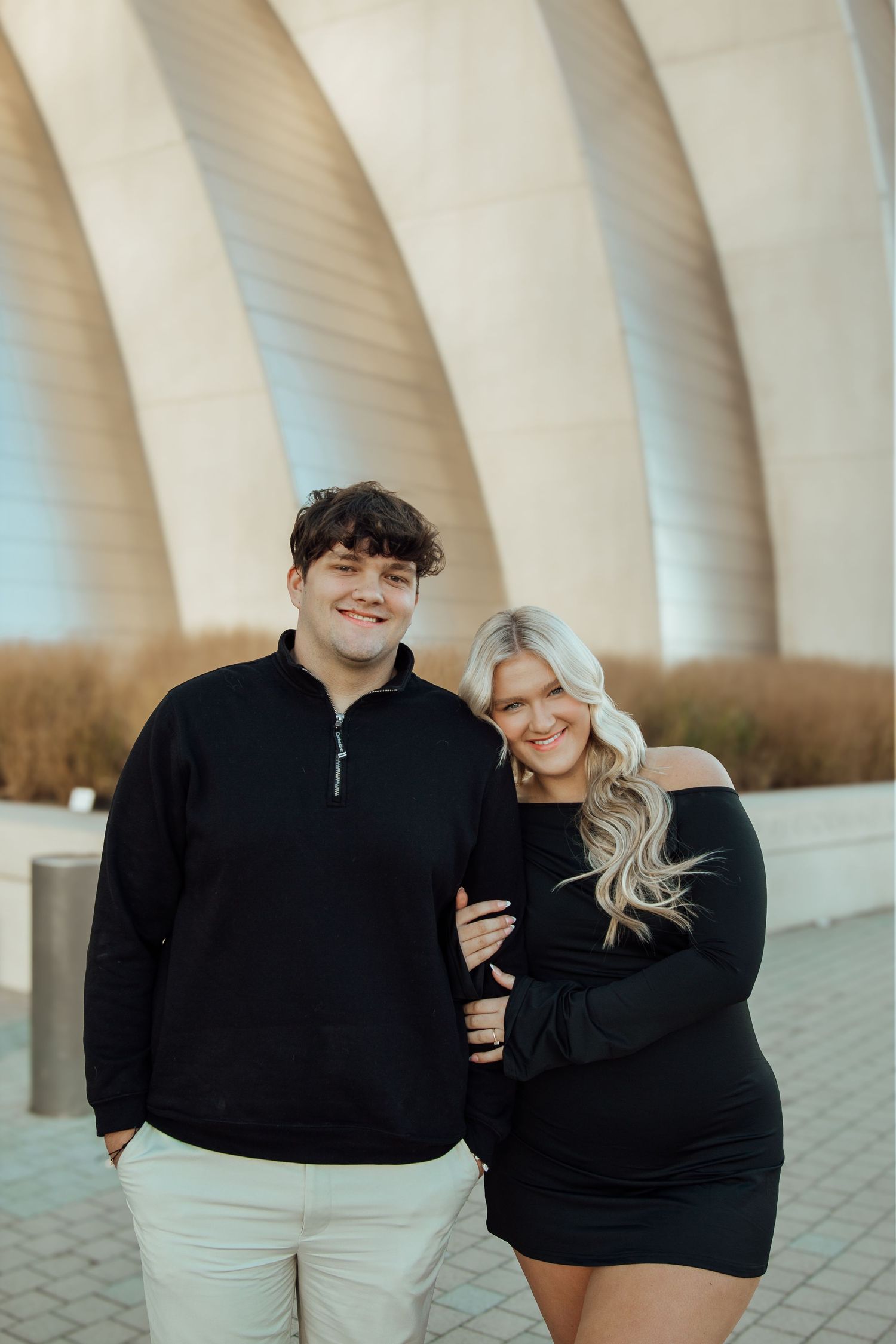 A couple in matching black outfits smiles together in front of a modern architectural structure outdoors.