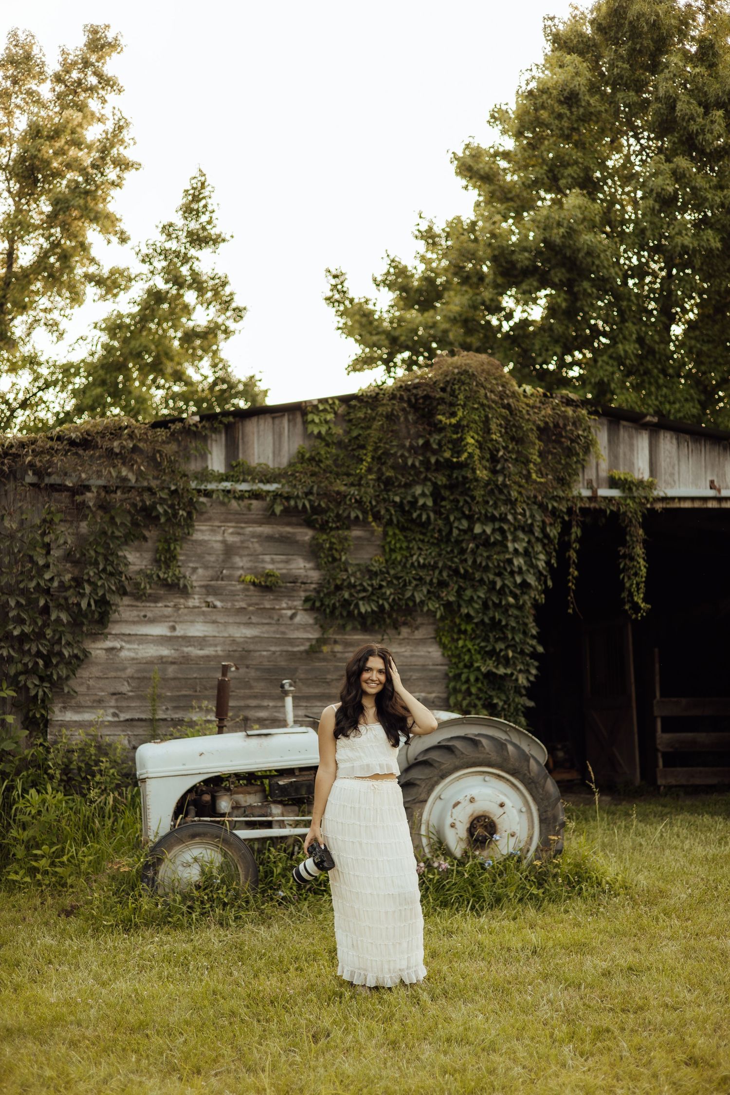 Woman in white lace dress standing in front of vintage tractor and ivy-covered barn on a farm.