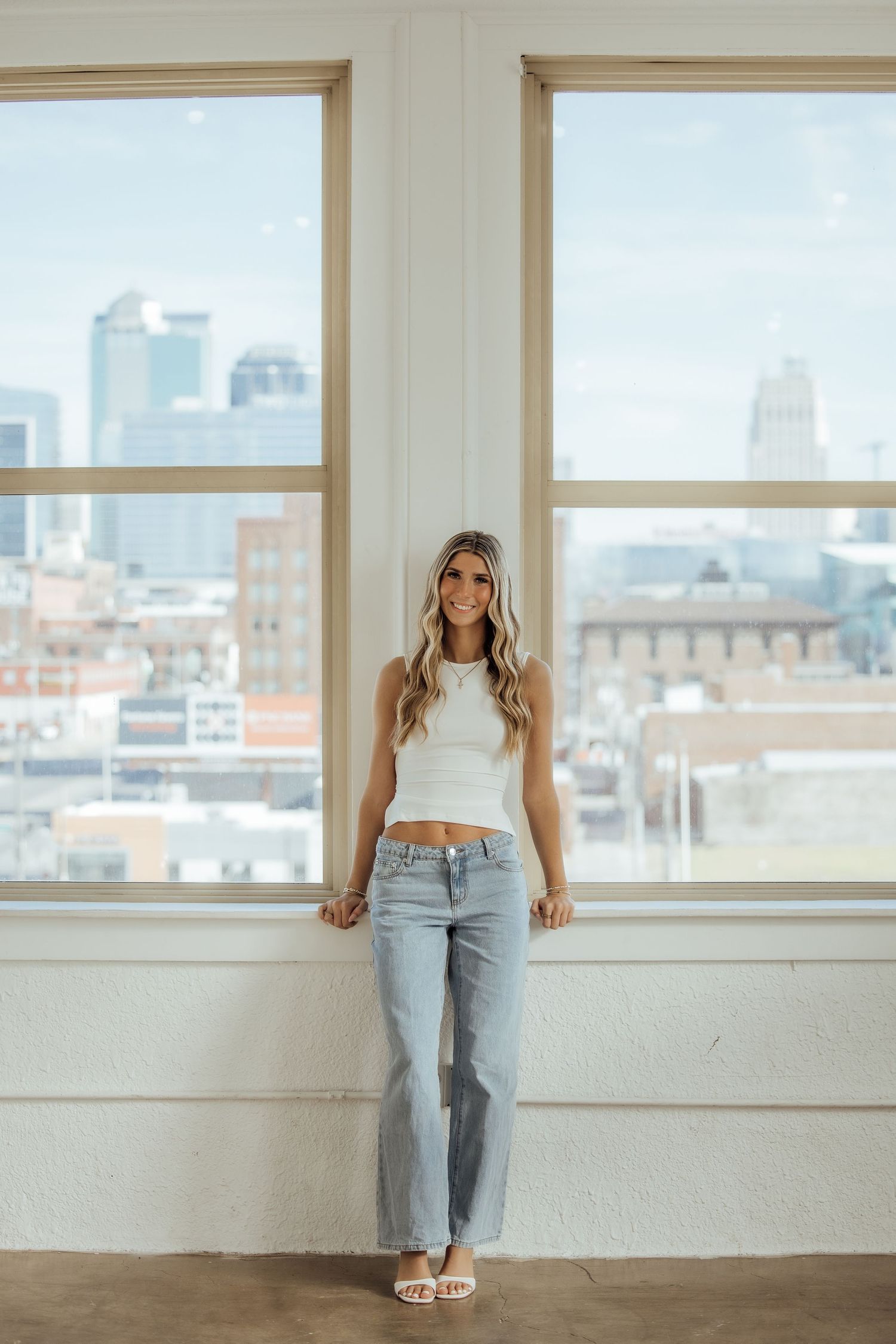 Smiling blonde woman in white crop top and jeans standing by large windows with city skyline.