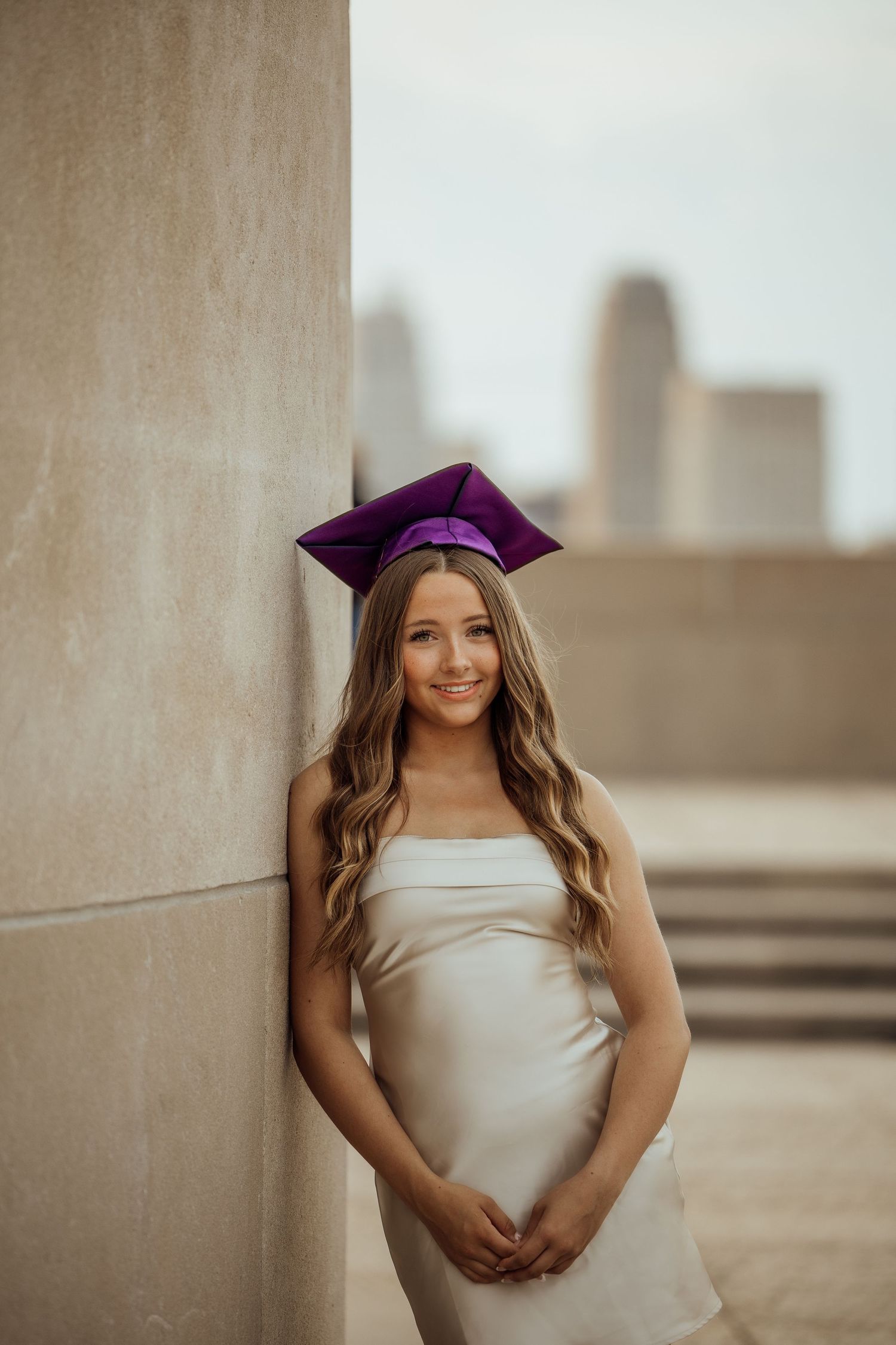 Smiling graduate in white dress and purple mortarboard cap leaning against stone column with city skyline.