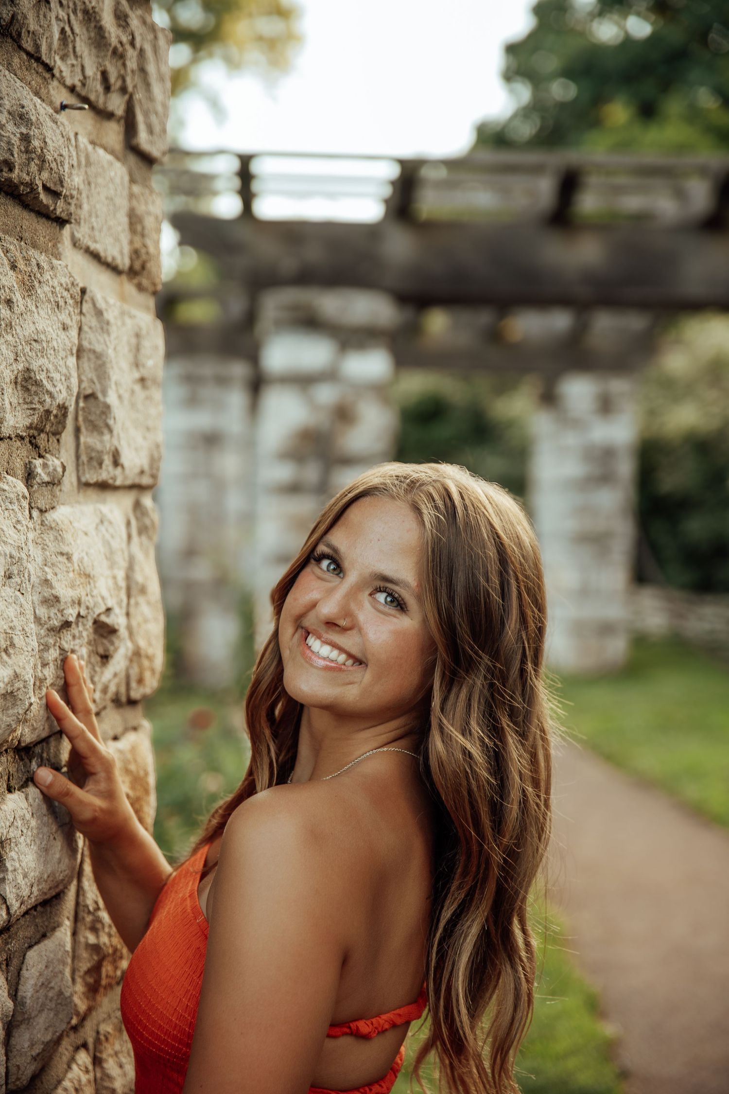 Smiling brunette woman in orange top touching rustic stone wall in garden with pergola background.