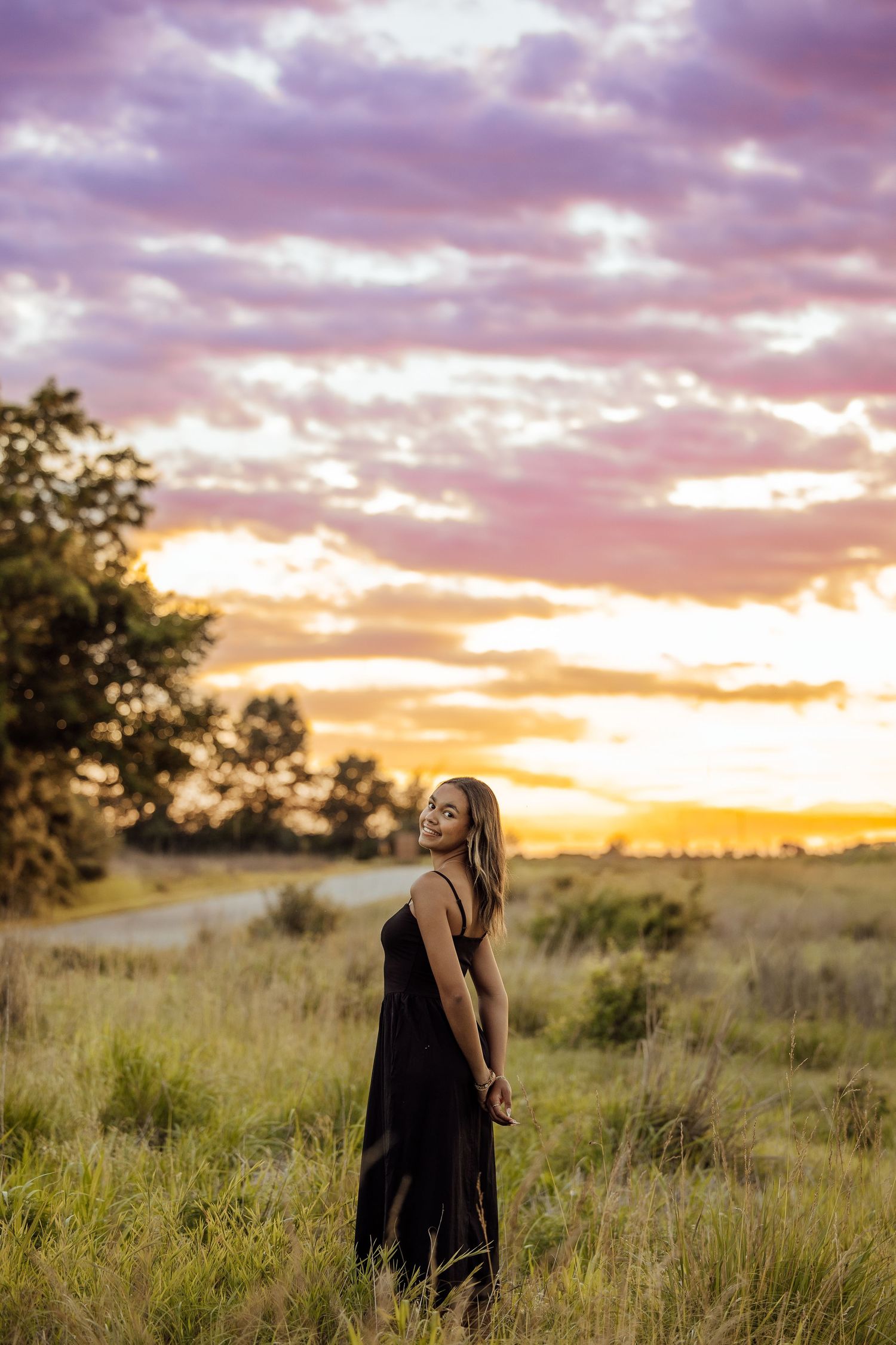 Woman in black dress standing in grassy field at golden sunset with dramatic purple and orange sky.