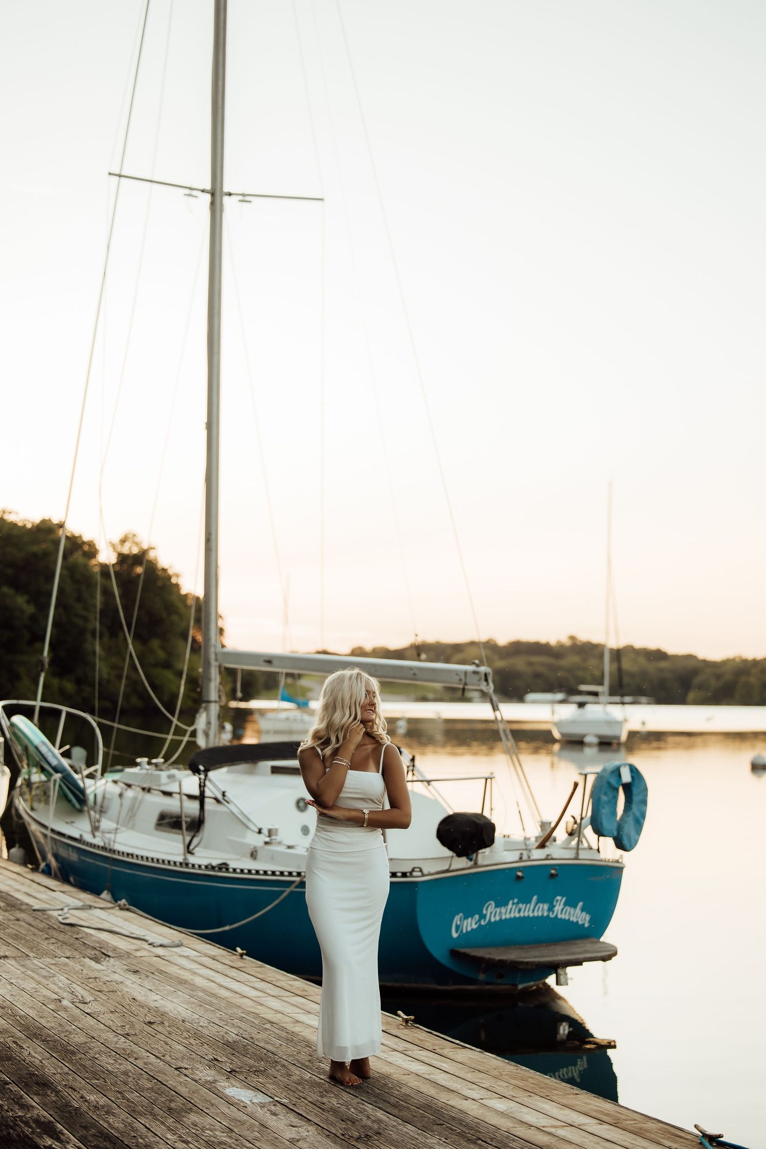 Blonde woman in white dress standing on wooden dock beside teal sailboat at golden hour sunset.