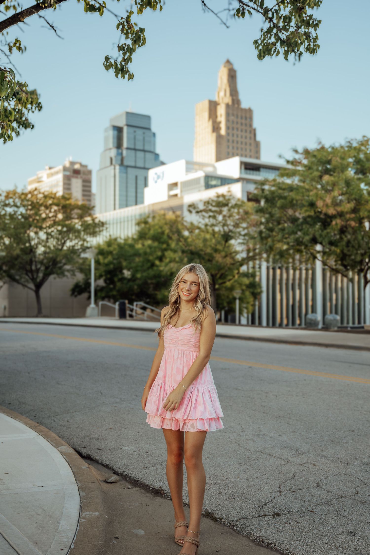 Smiling blonde woman in pink mini dress posing on urban street with city skyline buildings in background.